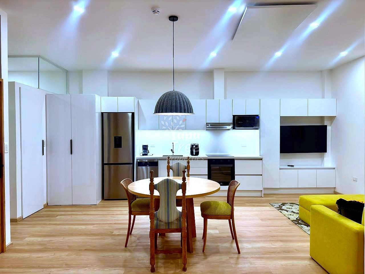 Open-plan departamento with white cabinetry, stainless steel appliances, round wood dining table with green chairs, and yellow sofa on wood flooring.