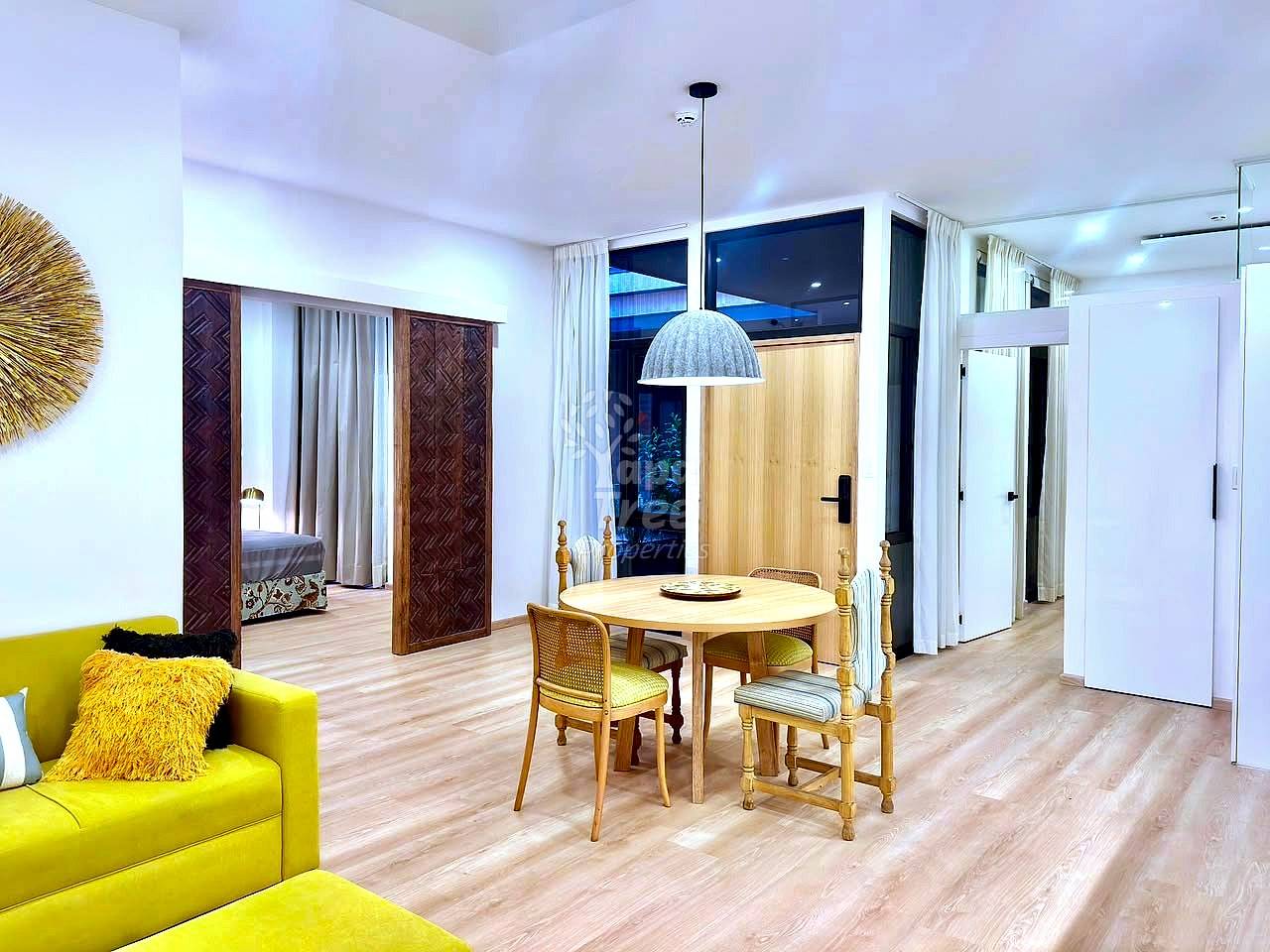 Open-plan departamento with wooden dining table, yellow sofa, wooden chairs, pendant light, wood flooring, and glass doors opening to bedroom area.