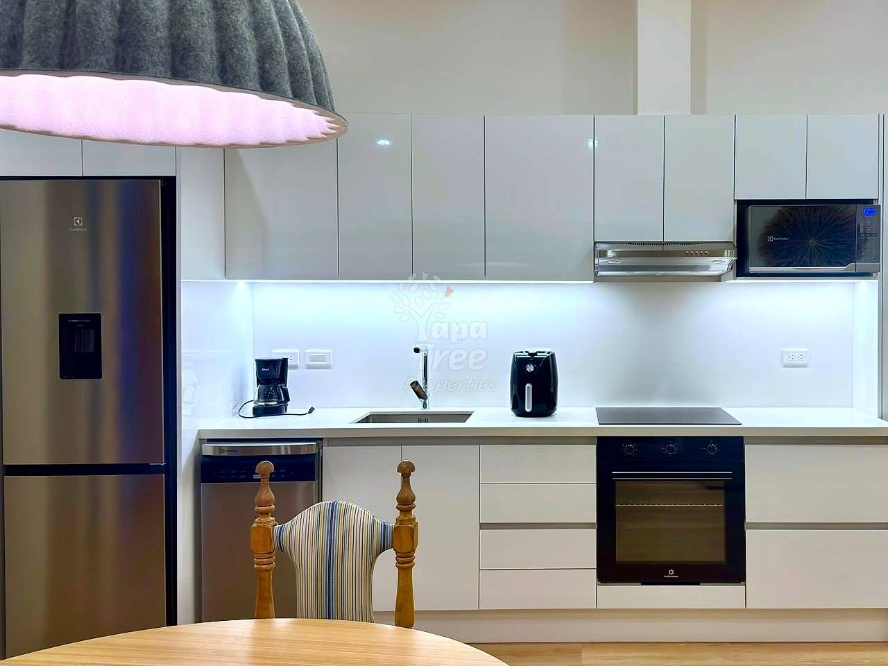 Modern kitchen with white cabinetry, stainless steel refrigerator, black electric oven, sink with faucet, and countertop appliances under pendant lighting.