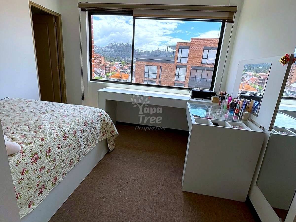 Bedroom with floral patterned bed, brown carpet floor, white desk, and large windows overlooking Cuenca cityscape with mountains and brick buildings.