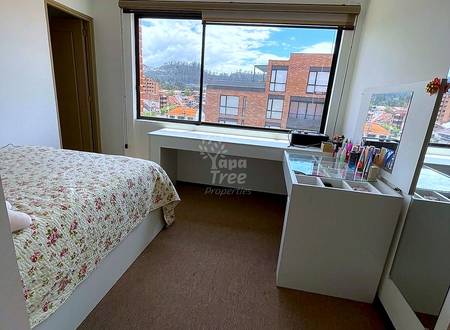 Bedroom with floral patterned bed, brown carpet floor, white desk, and large windows overlooking Cuenca cityscape with mountains and brick buildings.