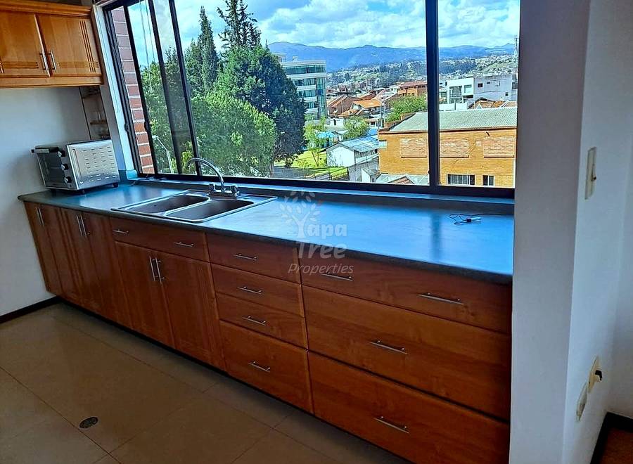 Modern cocina with wooden cabinetry, blue countertop, stainless steel sink, and large windows overlooking Cuenca cityscape and mountains.