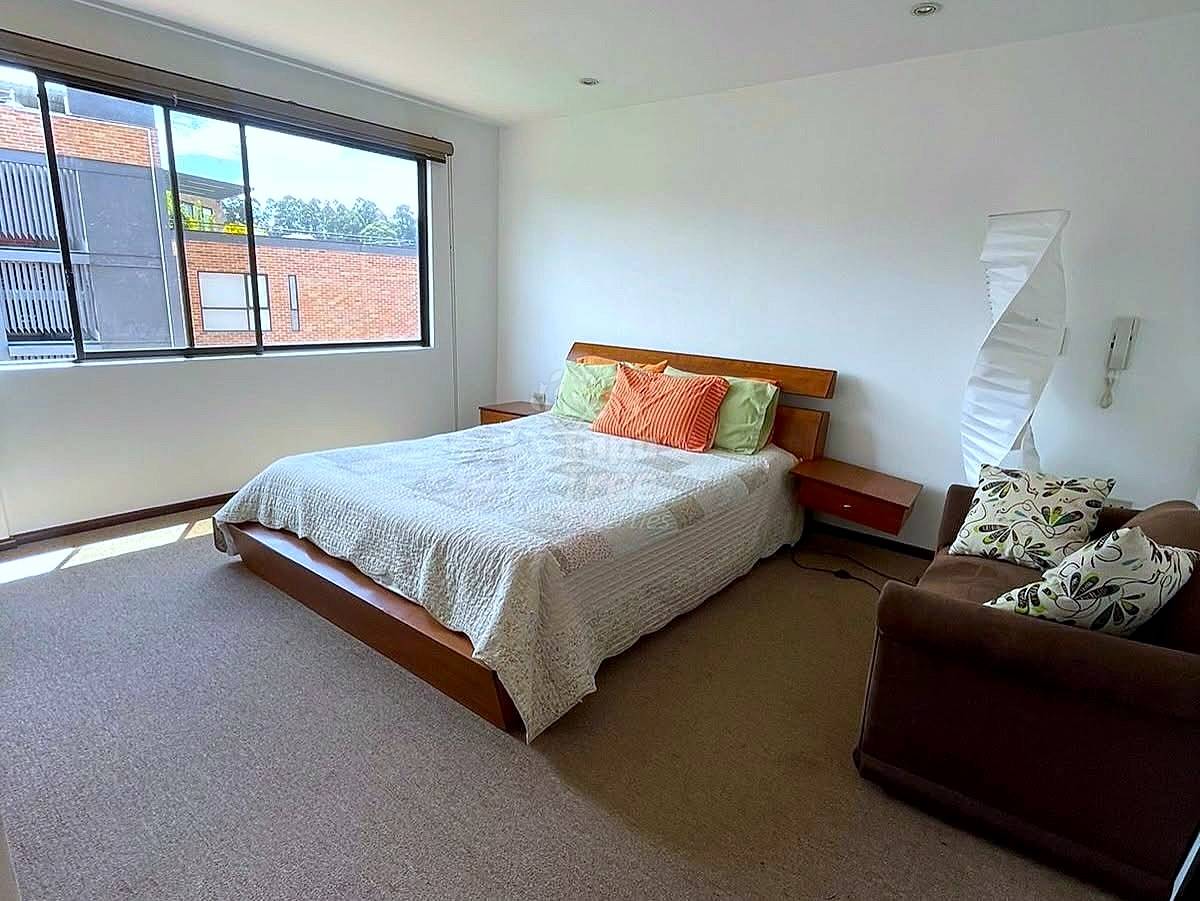 Bright bedroom with wooden bed, gray carpet, brown sofa, large window overlooking brick building, and white walls.