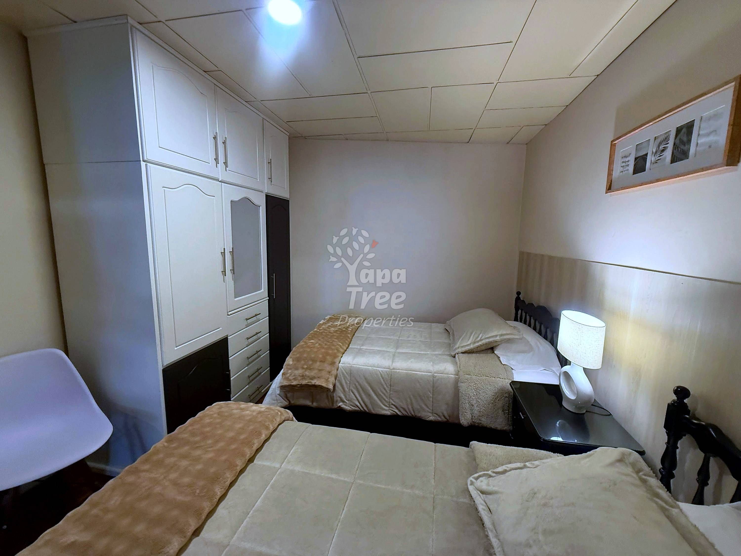 Small bedroom with two single beds, white built-in wardrobe, ceiling lights, and beige walls with wainscoting.