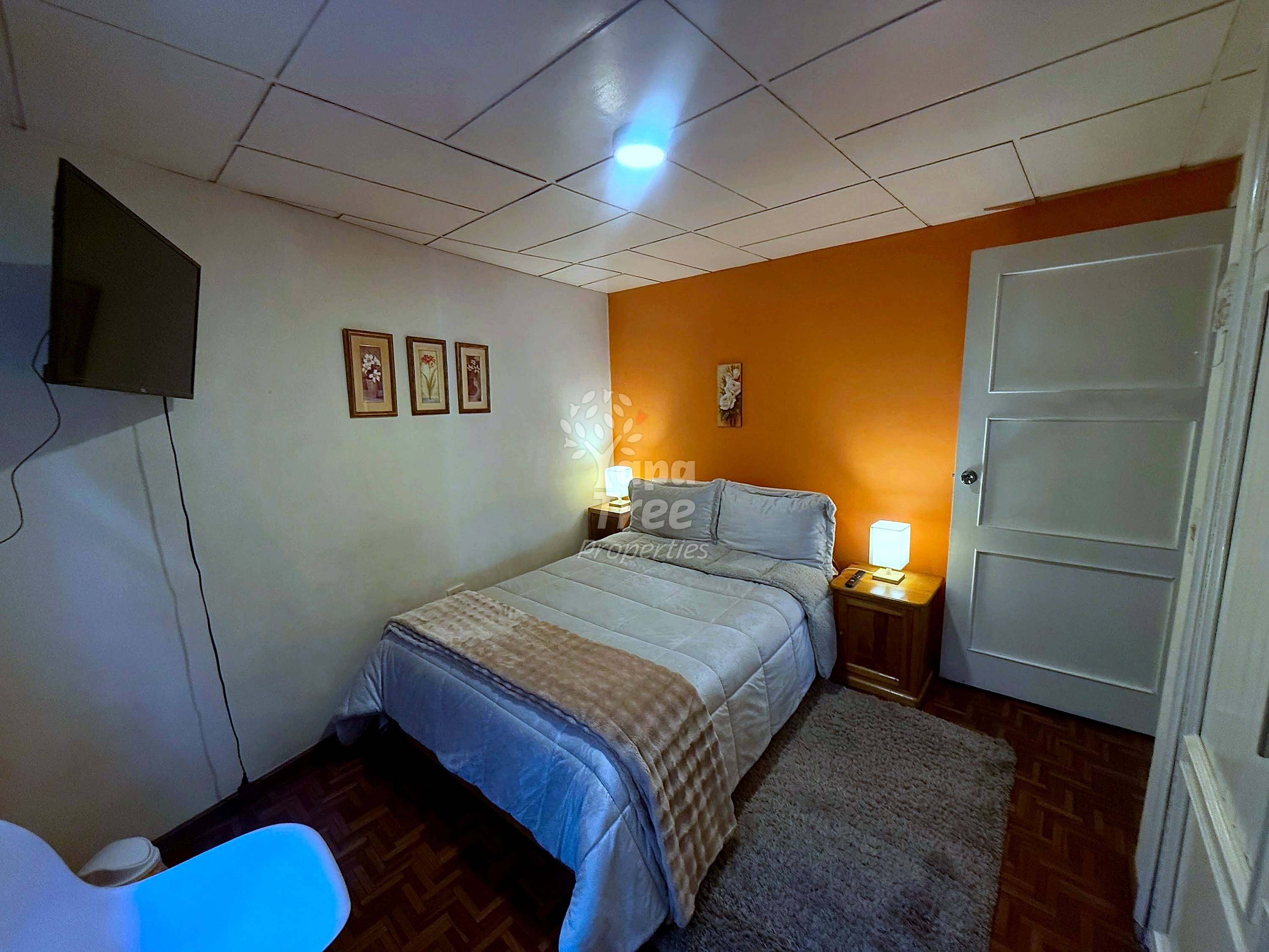 Bedroom with queen bed, orange accent wall, drop ceiling, wall-mounted TV, wooden nightstands with lamps, and white closet door.