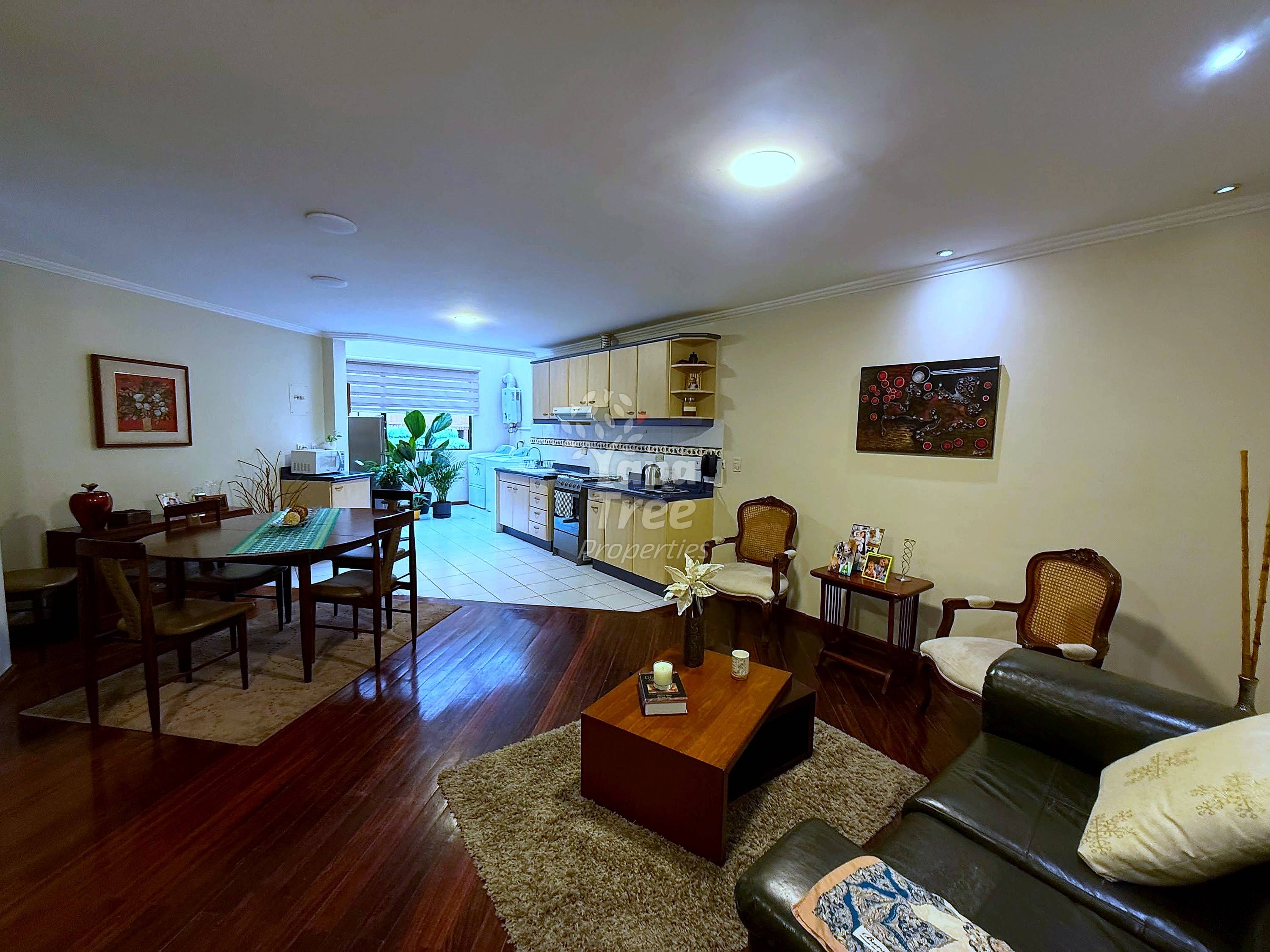 Open-plan living room and kitchen with dark wood flooring, beige walls, recessed lighting, dining table, black leather sofa, and equipped cocina with wooden cabinetry.