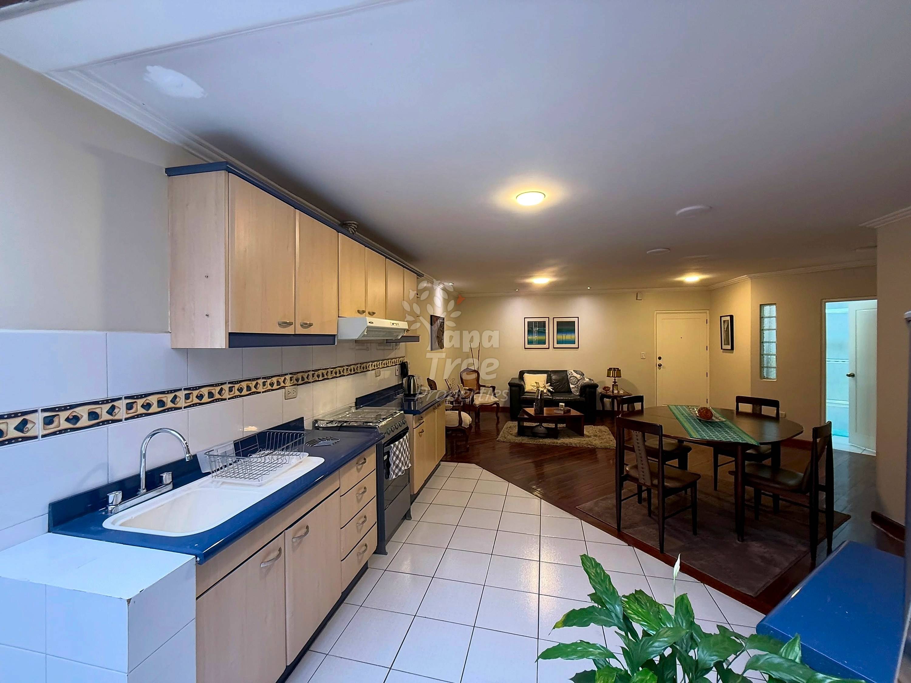 Open-plan cocina and dining area with light wood cabinets, blue countertops, gas stove, and ceramic tile floors opening to living room with dark wood furniture.
