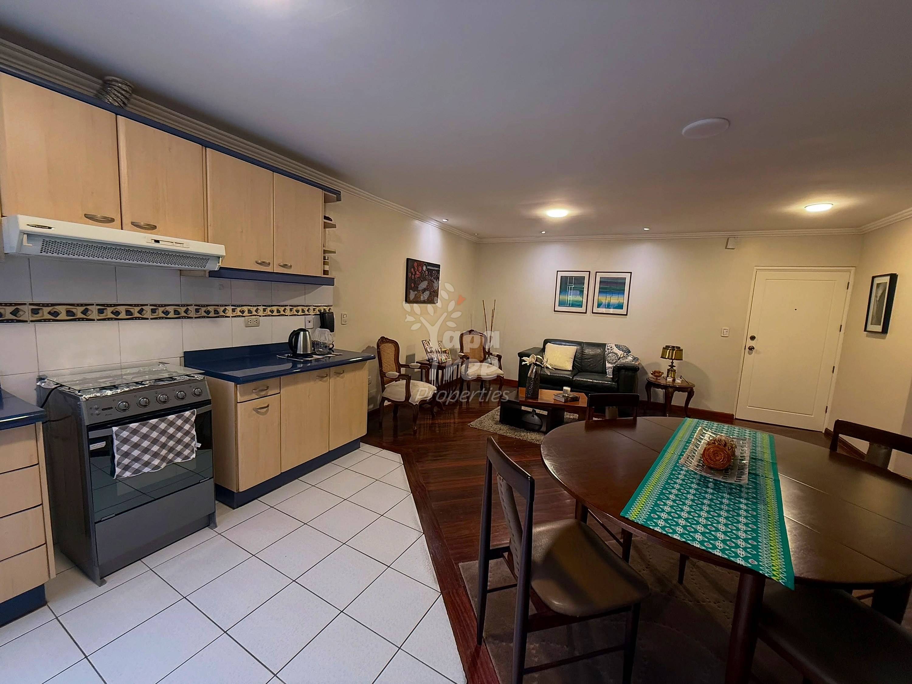 Open-plan kitchen and dining area with cream cabinets, black countertops, gas stove, dining table with wooden chairs, and living space beyond.