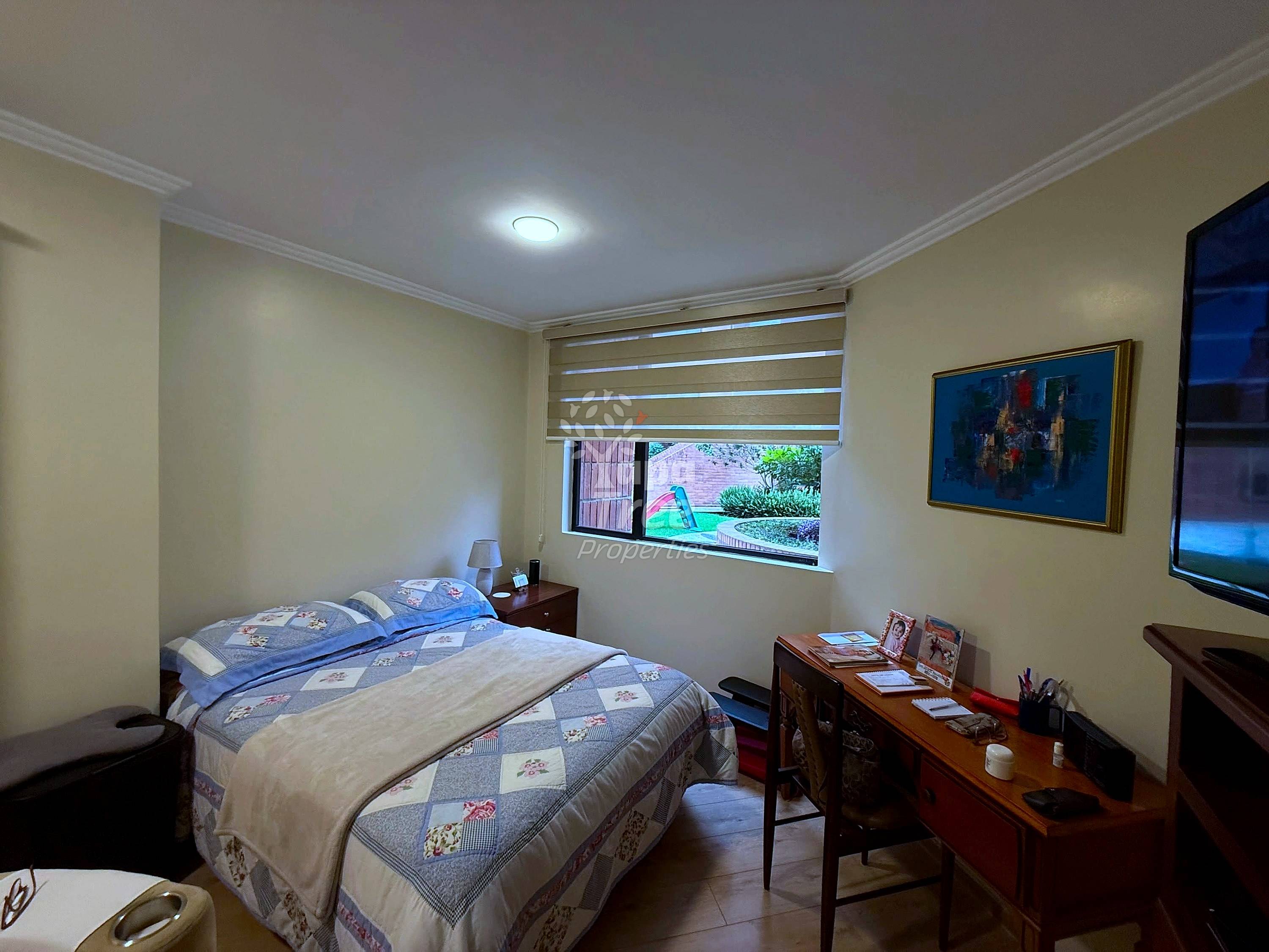 Bedroom with queen bed, patterned bedding, wooden nightstand, desk with chair, TV mounted on wall, and window with horizontal blinds showing outdoor garden view.