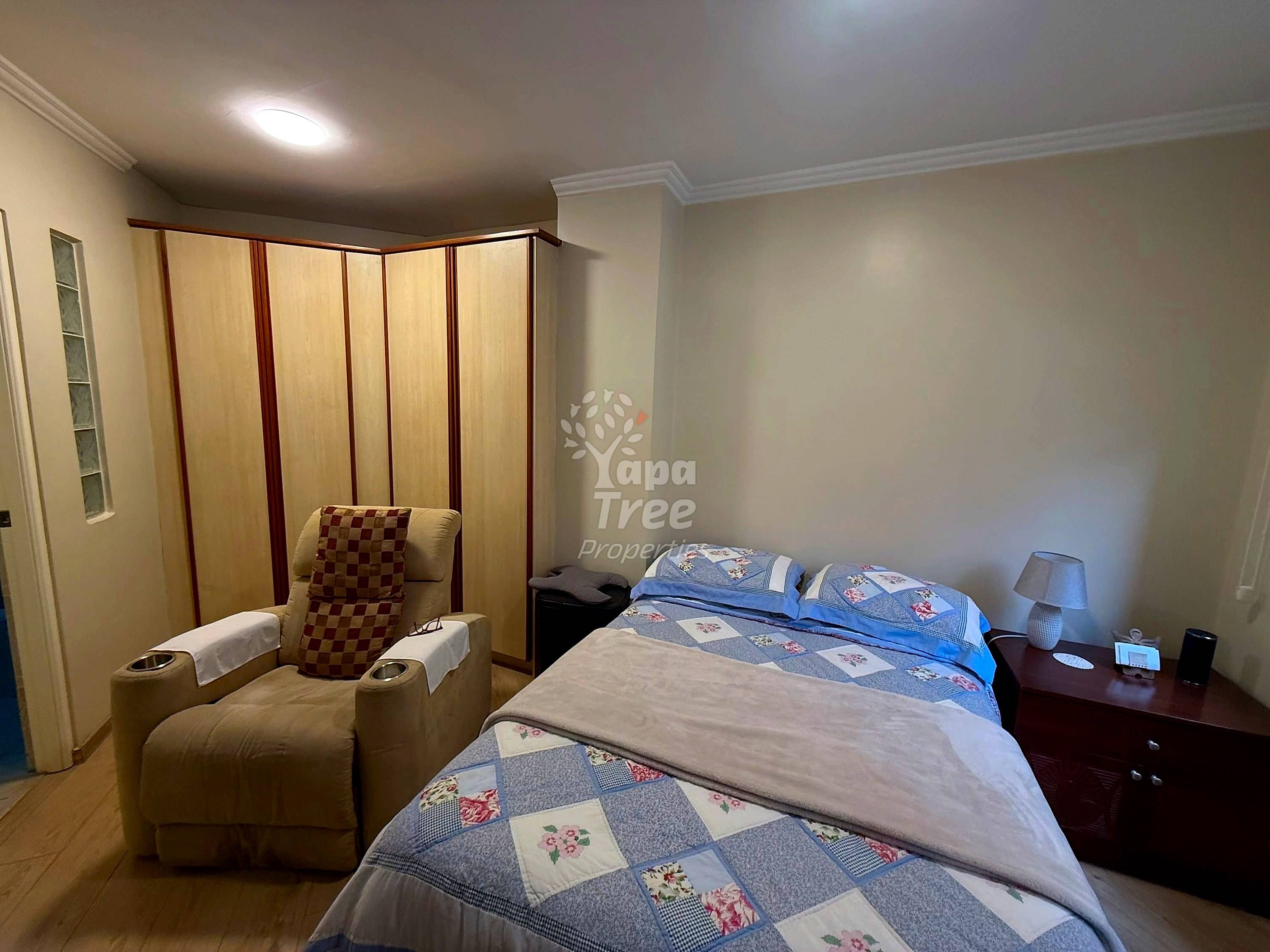 Bedroom with two beds, patchwork quilts, wooden nightstand with lamp, beige armchair, folding room divider, and recessed ceiling light.