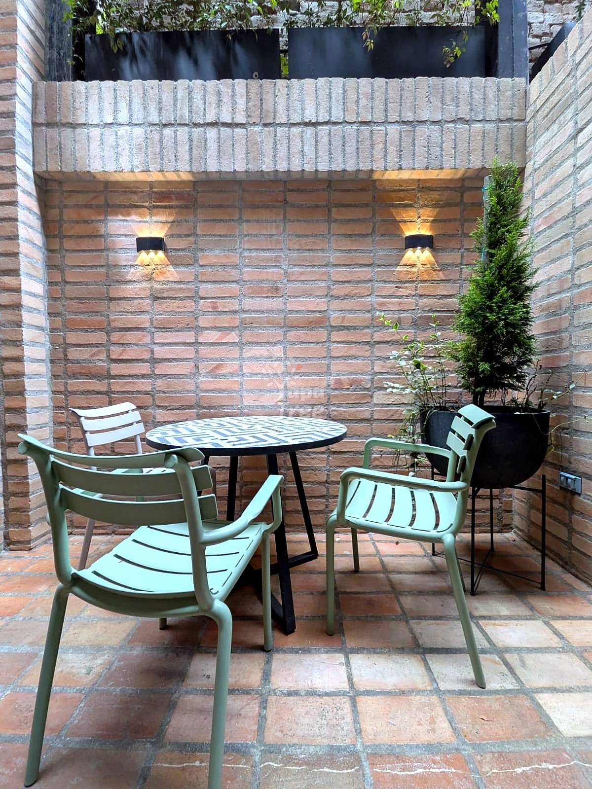 Brick patio with round table, four green chairs, terracotta floor, two wall sconces, and potted plants.