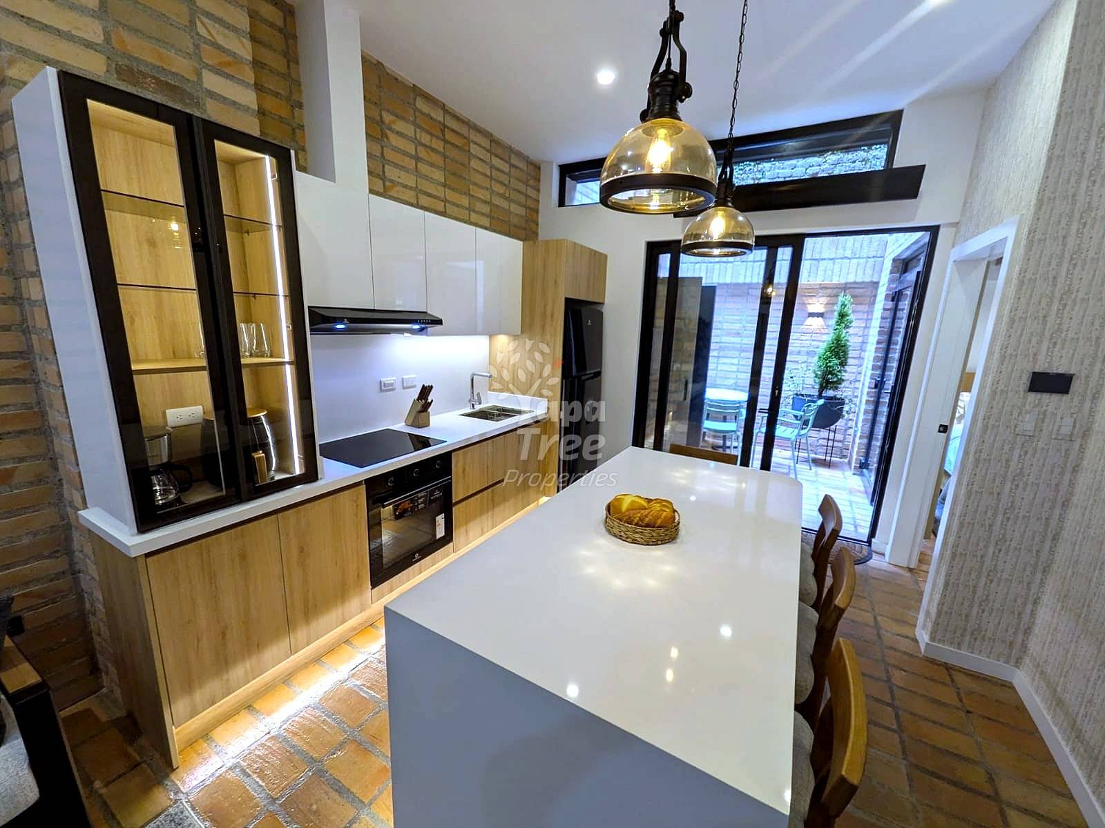 Modern kitchen with wood cabinetry, white island countertop, pendant lights, brick flooring, and glass doors opening to patio area.