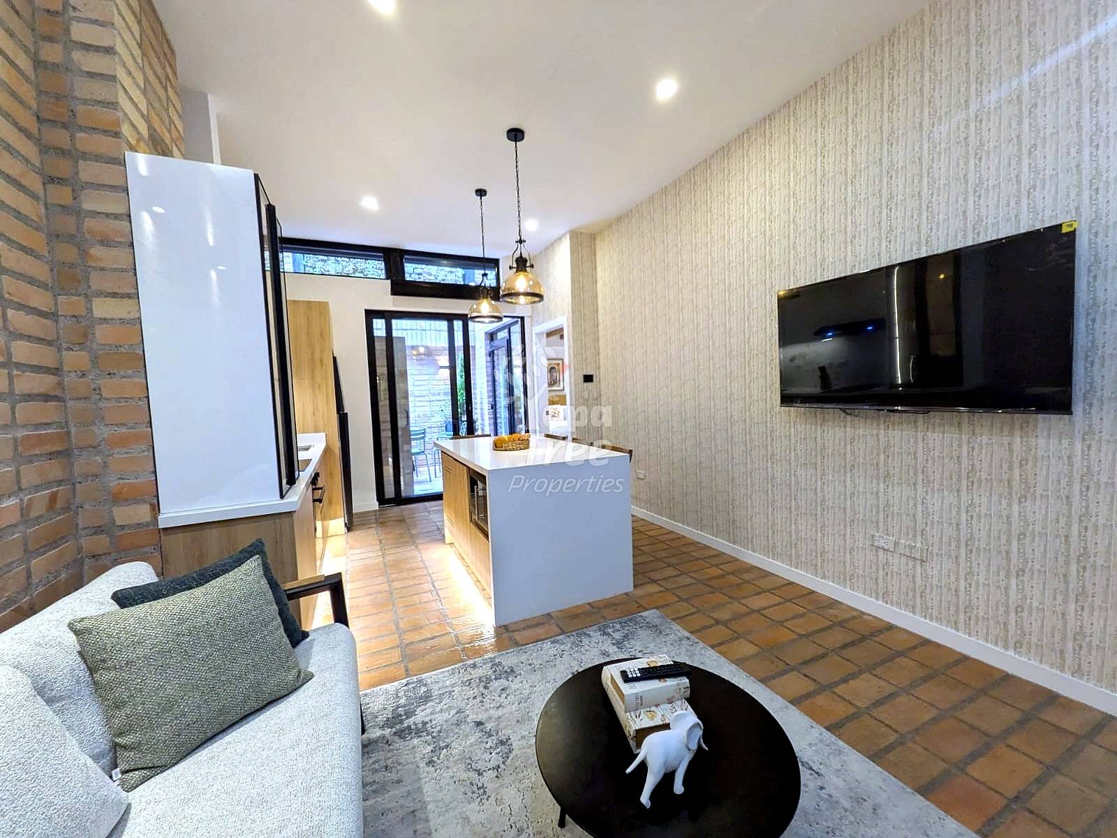 Open-concept living area with terracotta tile flooring, beige textured walls, white refrigerator, central kitchen island with pendant lighting, wall-mounted flat-screen TV, and upholstered seating.