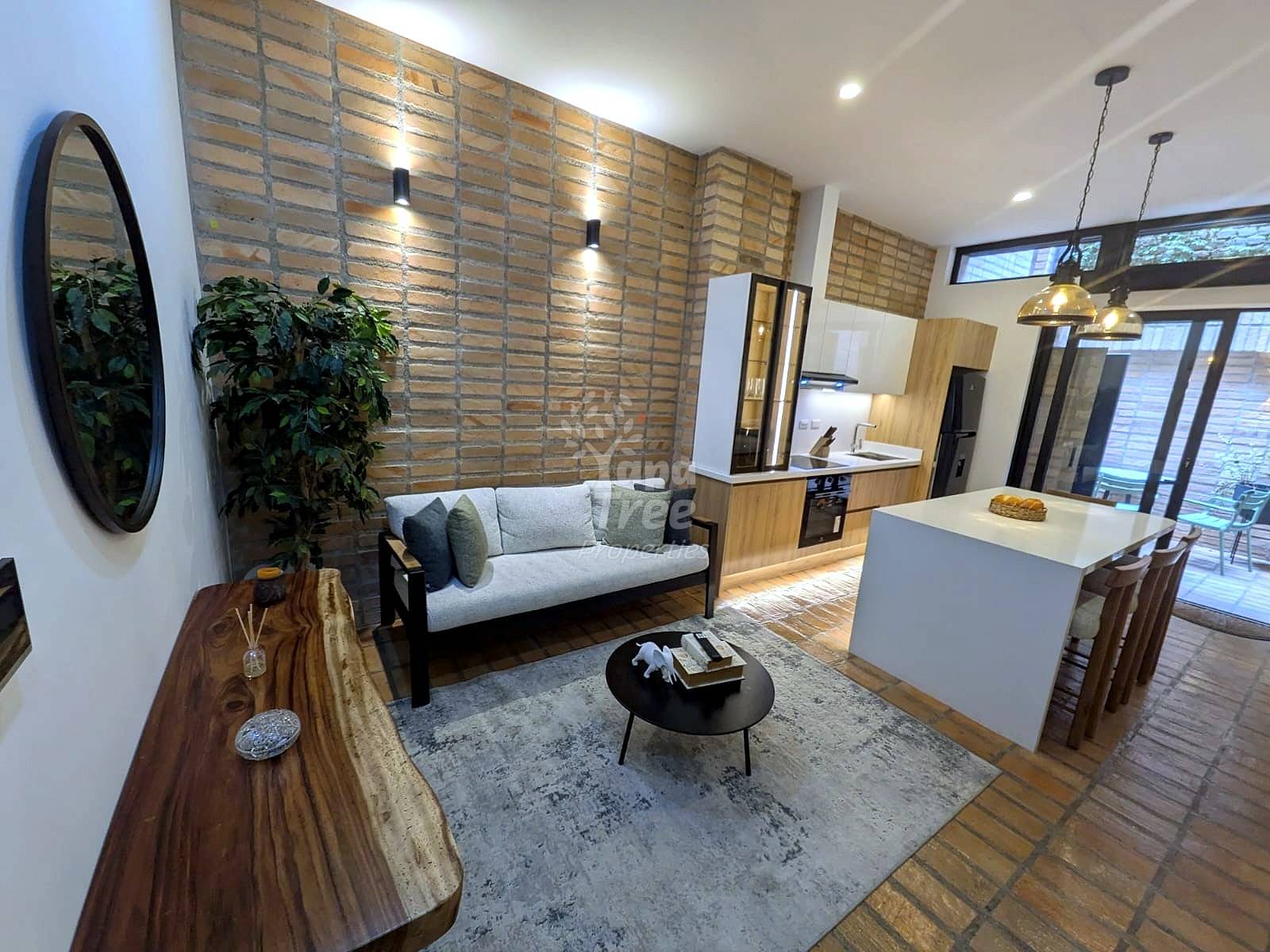 Open-concept departamento with living room, gray sofa, brick accent wall, pendant lights, white kitchen island, wood cabinetry, and glass doors to patio.