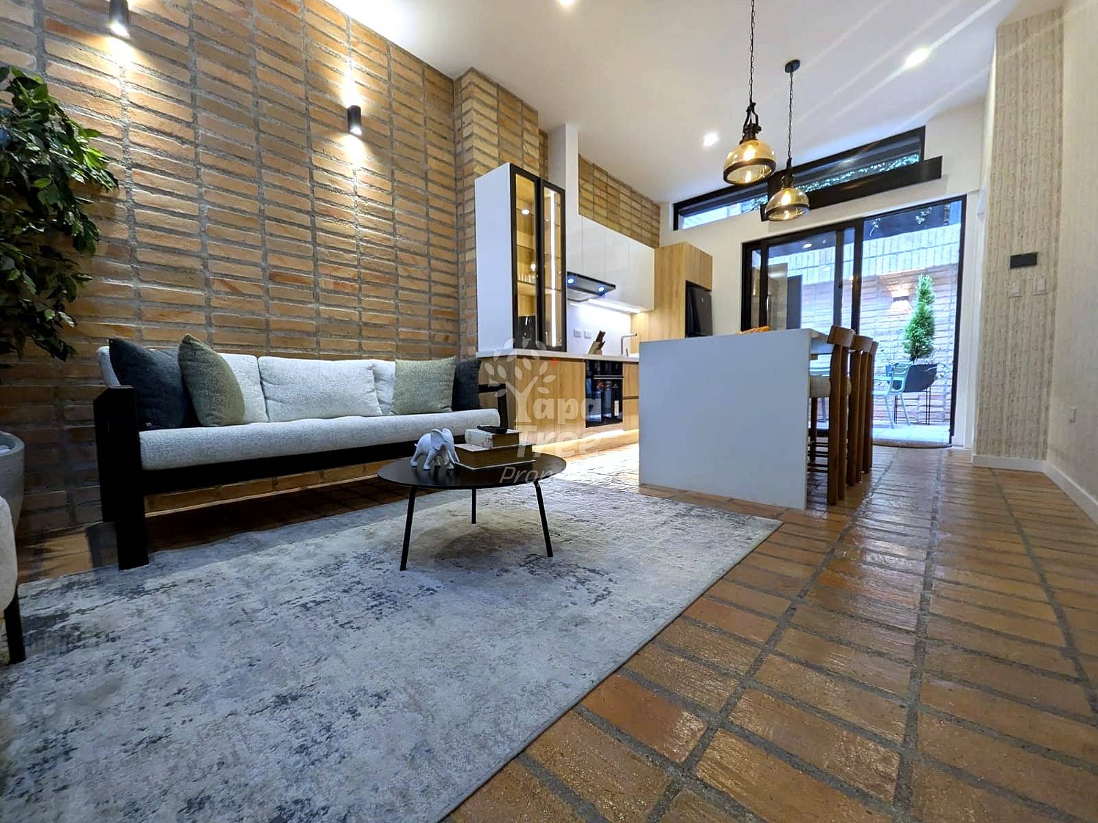 Open-plan living room with gray sofa, coffee table, exposed brick walls, pendant lighting, and terracotta tile flooring.