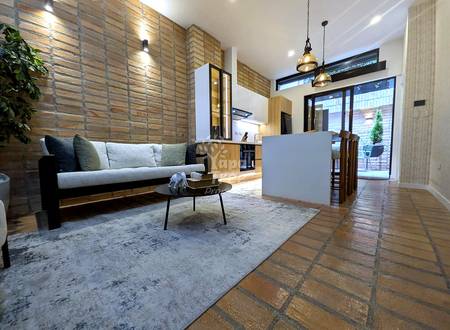 Open-plan living room with gray sofa, coffee table, exposed brick walls, pendant lighting, and terracotta tile flooring.