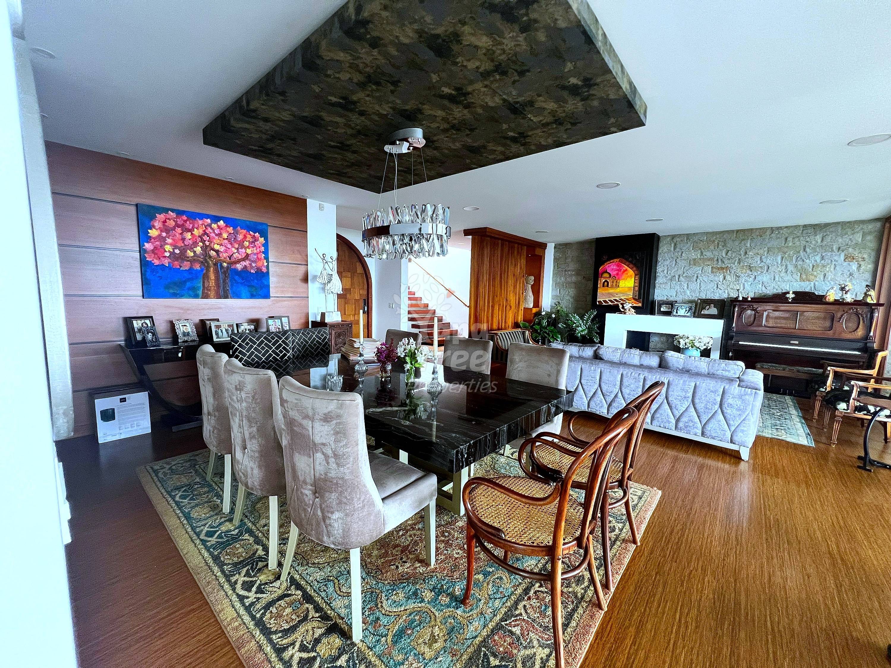 Open-concept living room with dining table, upholstered seating, wood floors, fireplace, piano, and decorative ceiling feature with staircase visible.
