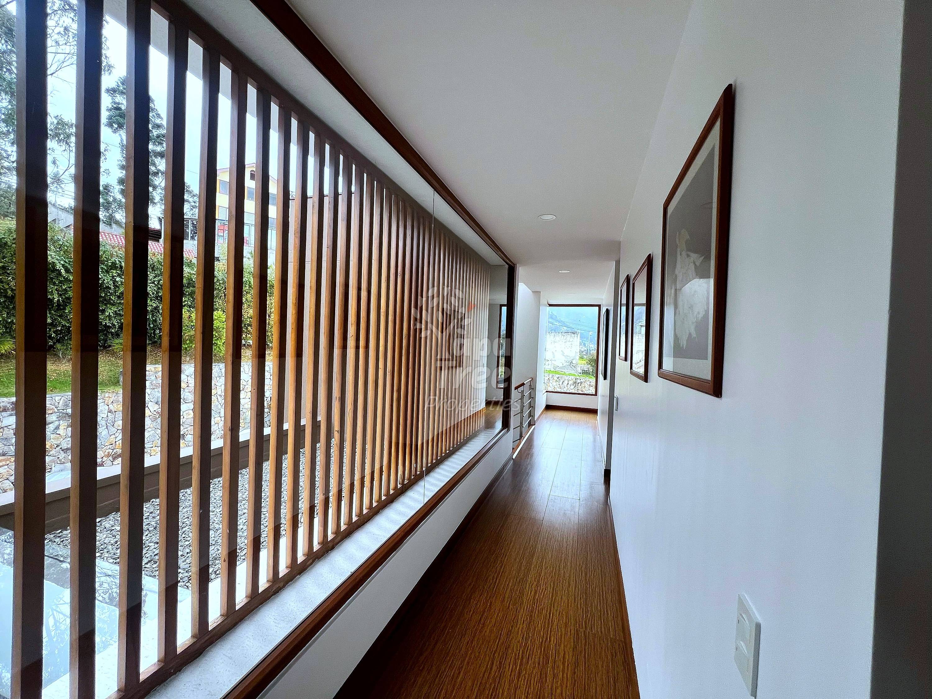 Hallway with dark wood flooring, white walls, vertical dark wood railing with glass panels on left side, framed artwork on right wall, natural light from windows.