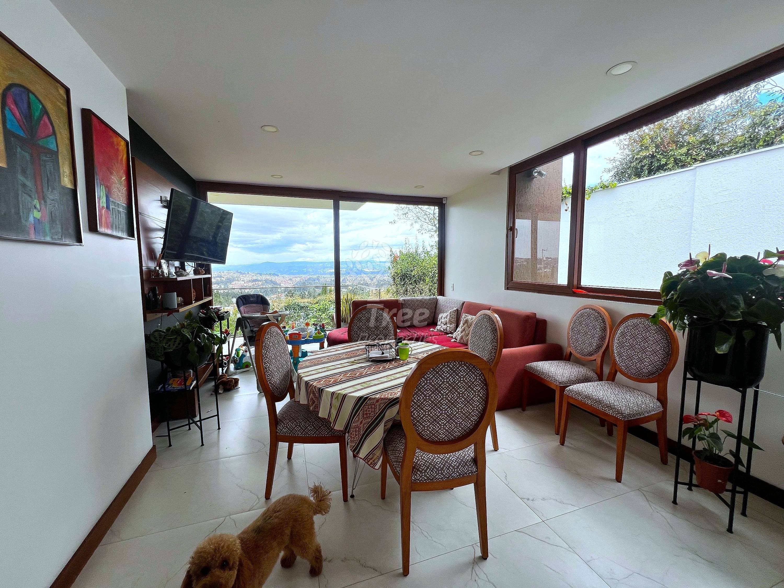 Living room with dining table, wooden chairs, red sofa, TV, large windows overlooking valley, recessed ceiling lights, tile flooring.