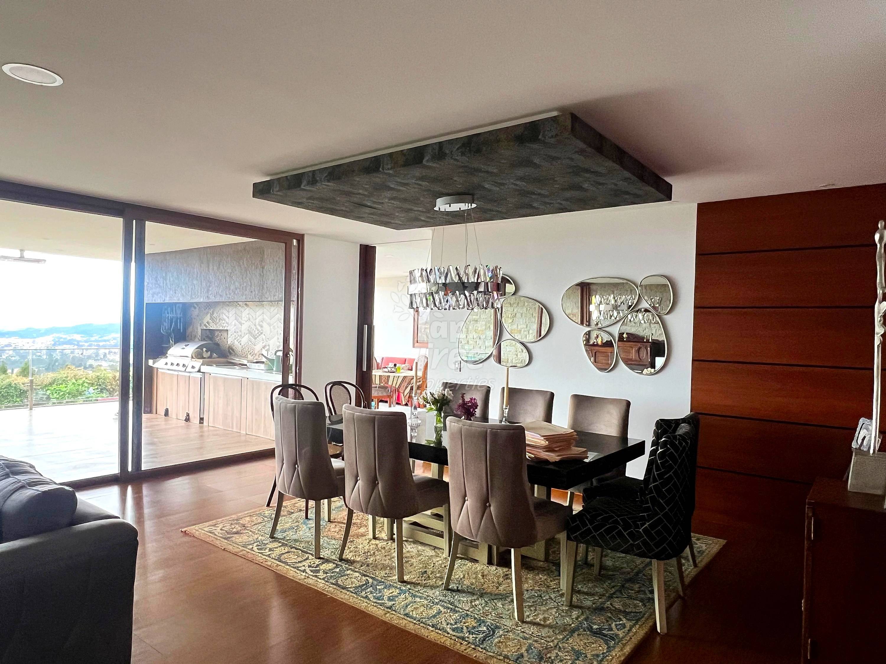 Dining room with black table, gray upholstered chairs, dark wood accent wall, recessed ceiling light fixture, and glass doors opening to terrace with valley views.