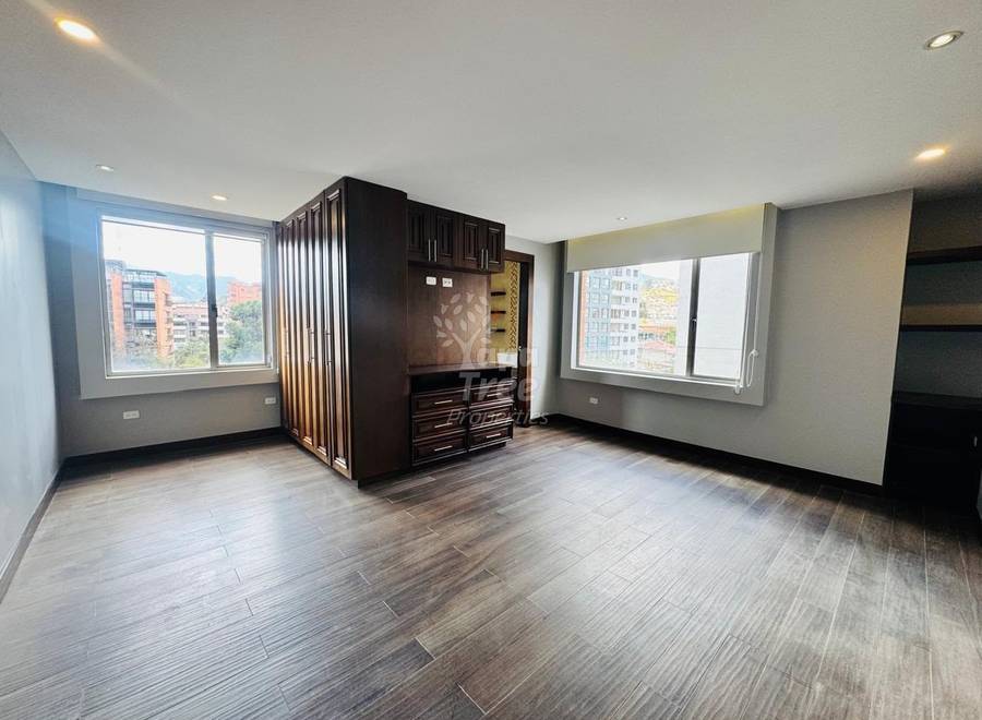 Bedroom with wood flooring, built-in dark wood closet with divider screen, two large windows with city views, recessed ceiling lights, and beige walls.