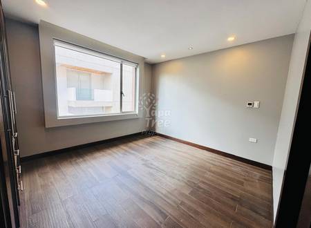 Empty room with wooden plank flooring, beige painted walls, recessed ceiling lights, and large window overlooking building exterior.