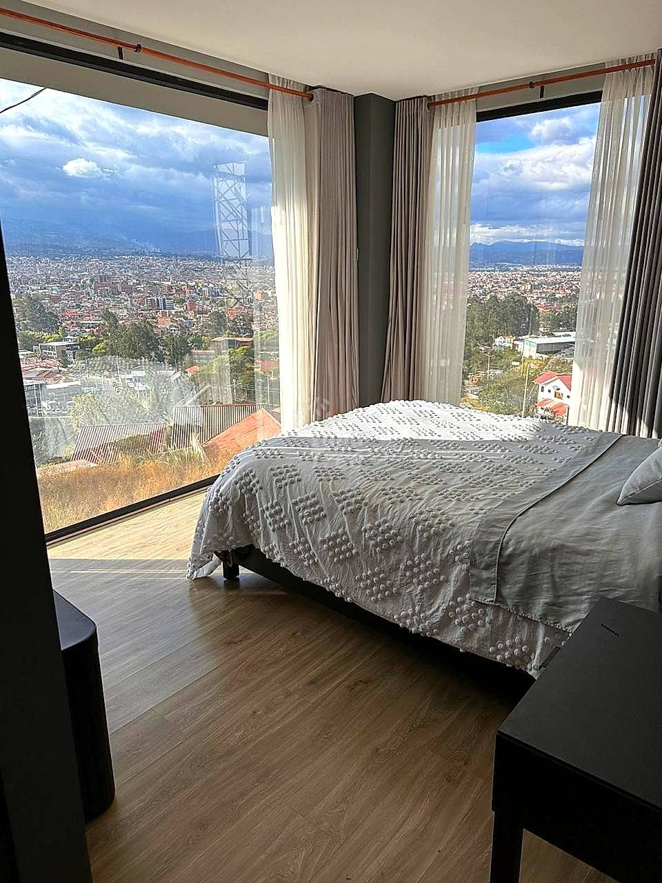 Bedroom with queen bed, gray bedding, wood flooring, floor-to-ceiling windows with sheer curtains overlooking Cuenca cityscape and mountains.