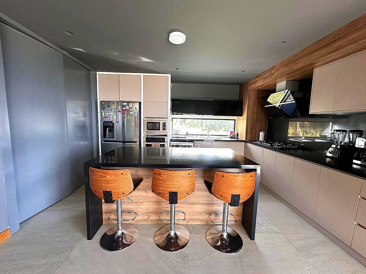 Cocina moderna con isla central oscura, tres taburetes de madera, electrodomésticos acero inoxidable, gabinetes blancos, y techo gris con iluminación empotrada.