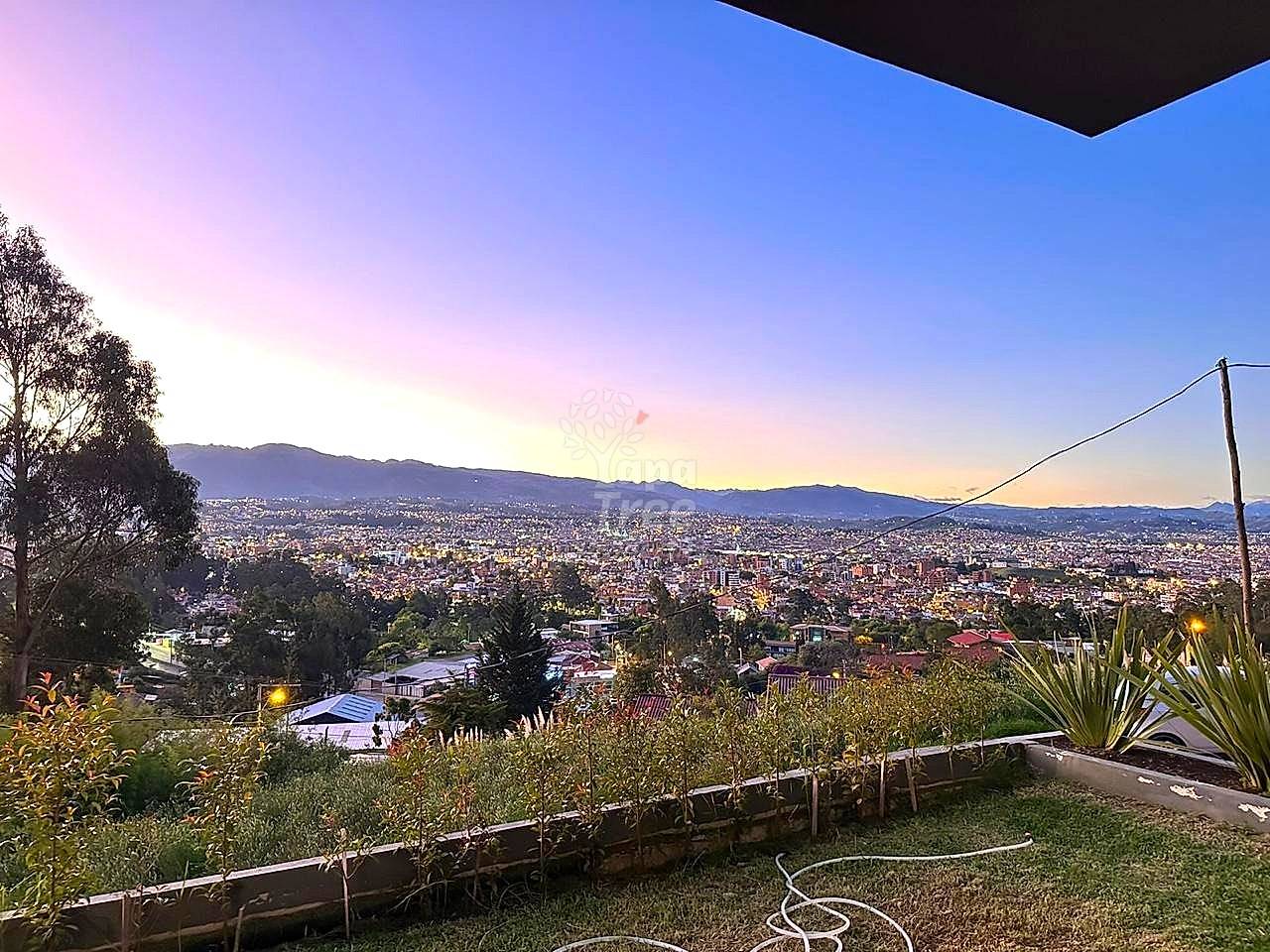 Elevated outdoor terrace with garden beds, plants, and power line, overlooking Cuenca city valley at dusk with mountains in background.