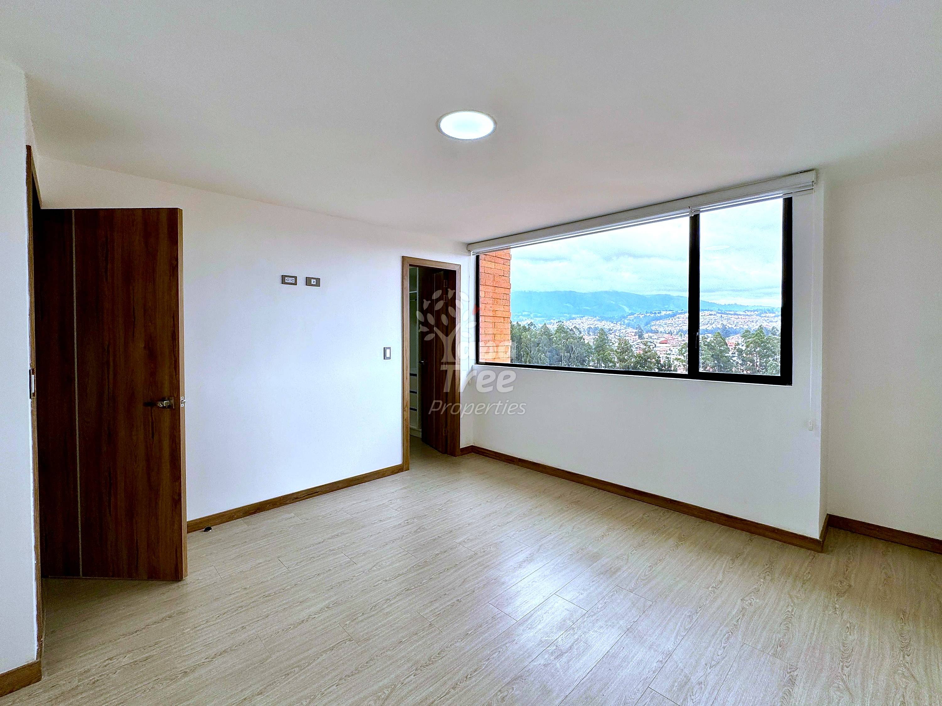 Empty room with laminate flooring, cream walls, dark wood doors, recessed ceiling light, and large window overlooking Cuenca cityscape and mountains.