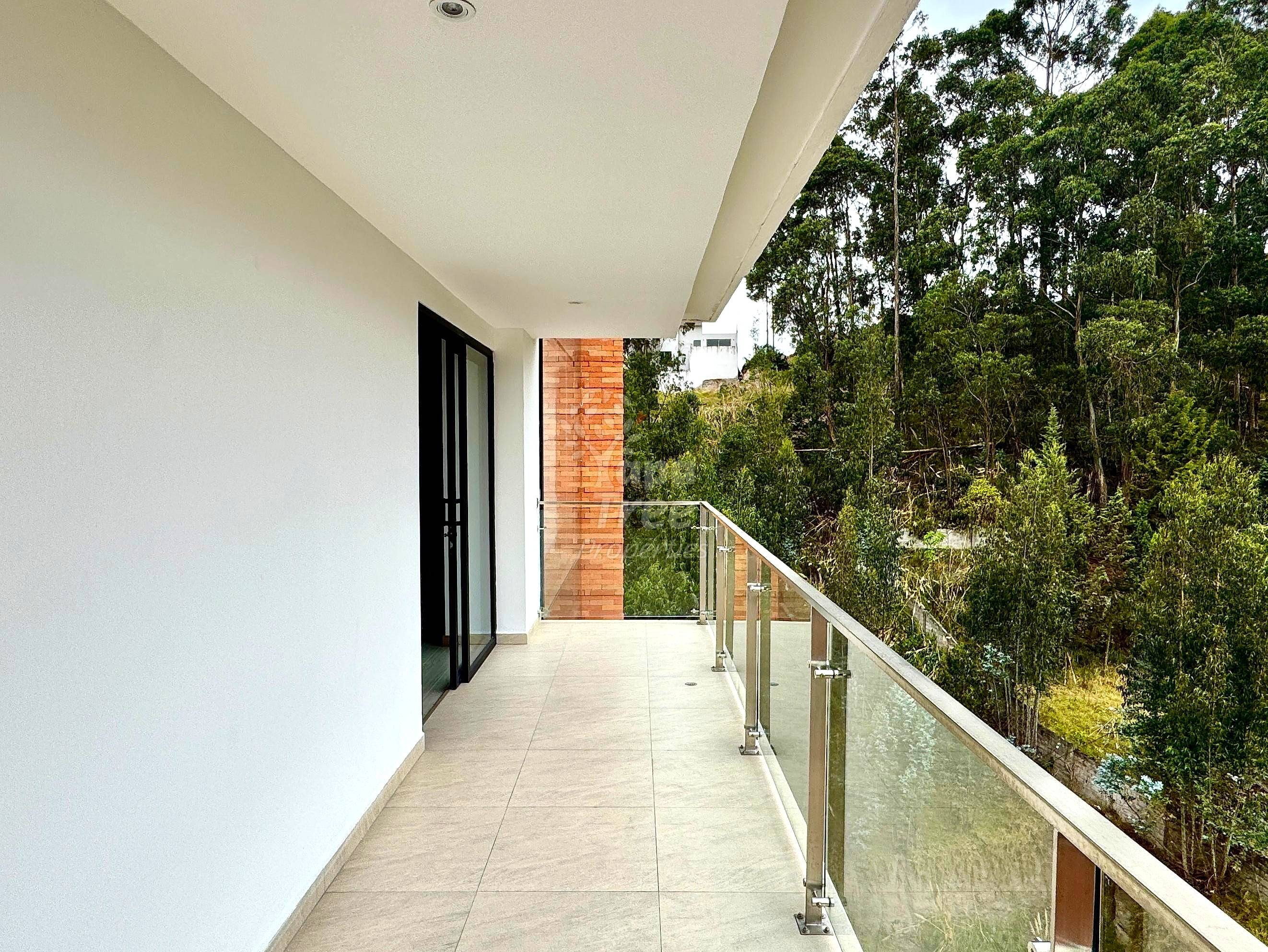 Covered balcony with light tile flooring, white walls, black glass doors, metal railing, and tree-lined hillside views.