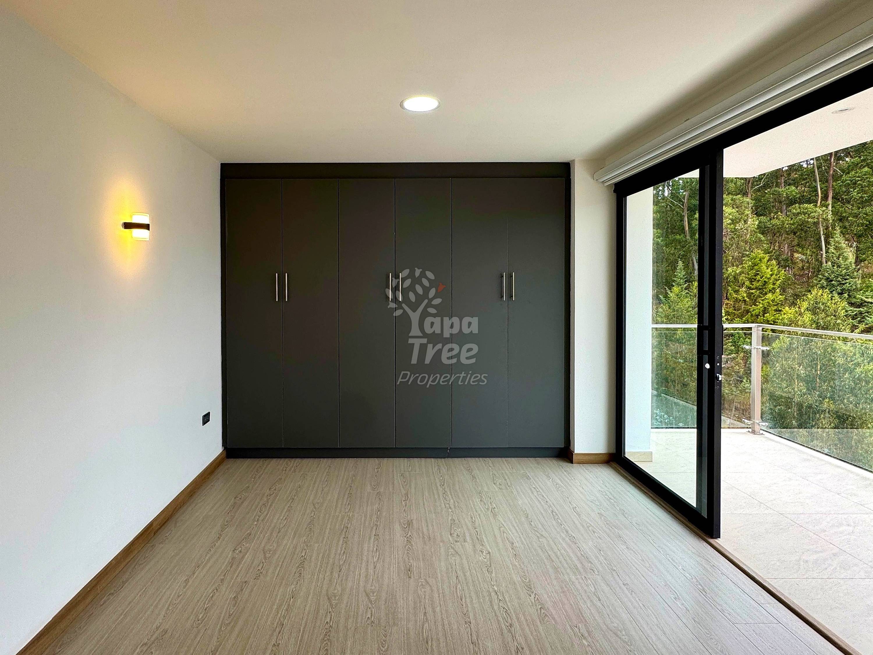 Bedroom with black six-door built-in wardrobe, wood-look flooring, white walls, recessed ceiling light, wall sconce, and glass door opening to forest balcony.
