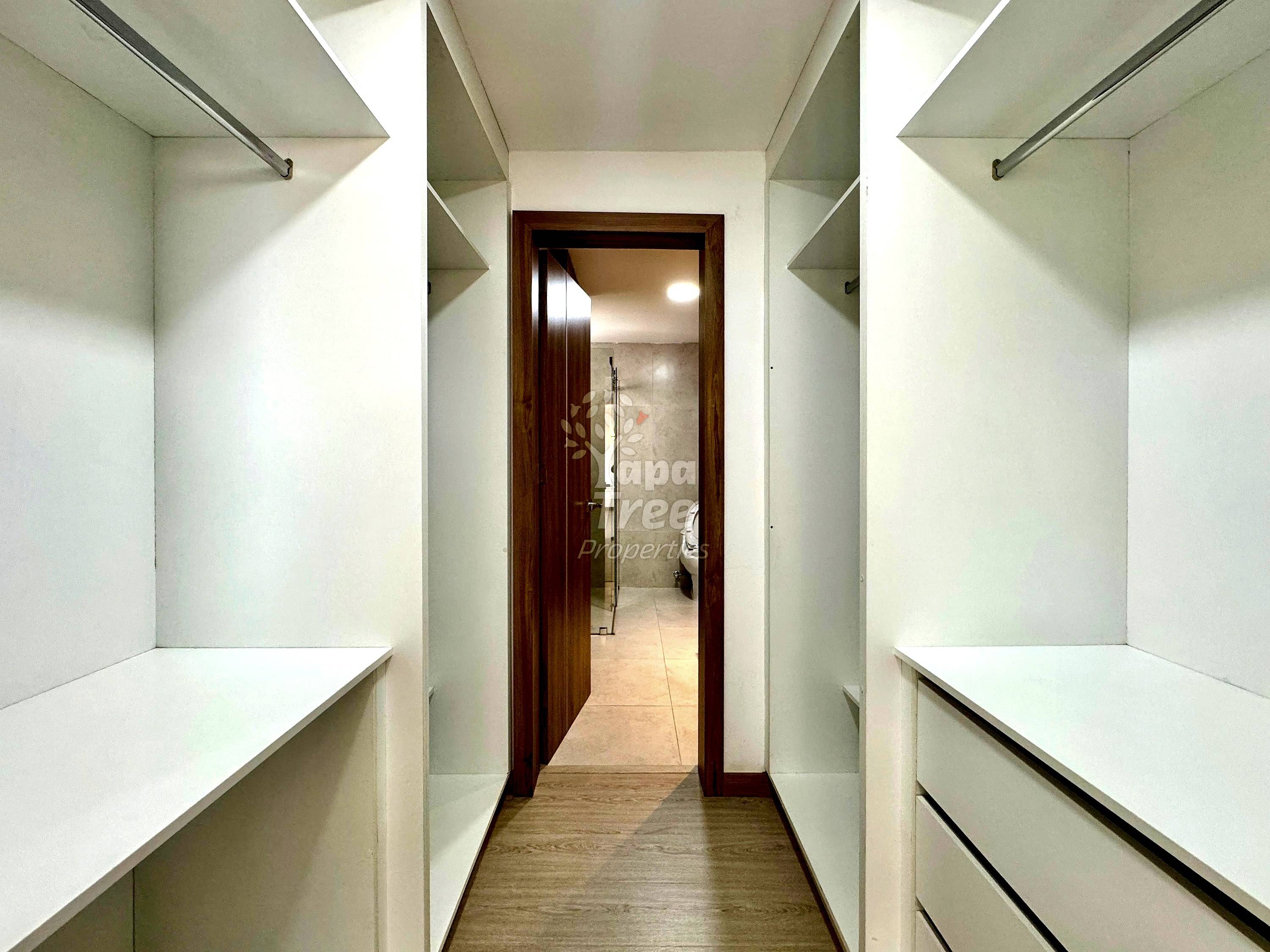 Walk-in closet with white built-in shelving on both sides, wood-framed doorway, and light wood flooring leading to bathroom.