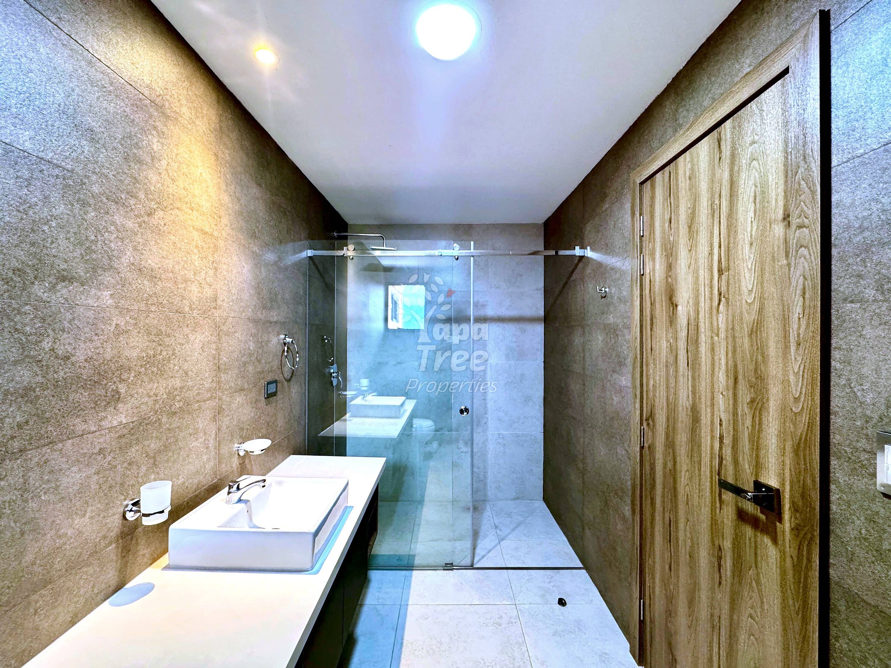 Modern bathroom with long vanity, white vessel sink, glass shower enclosure, beige tile walls, and wood-grain door on right side.