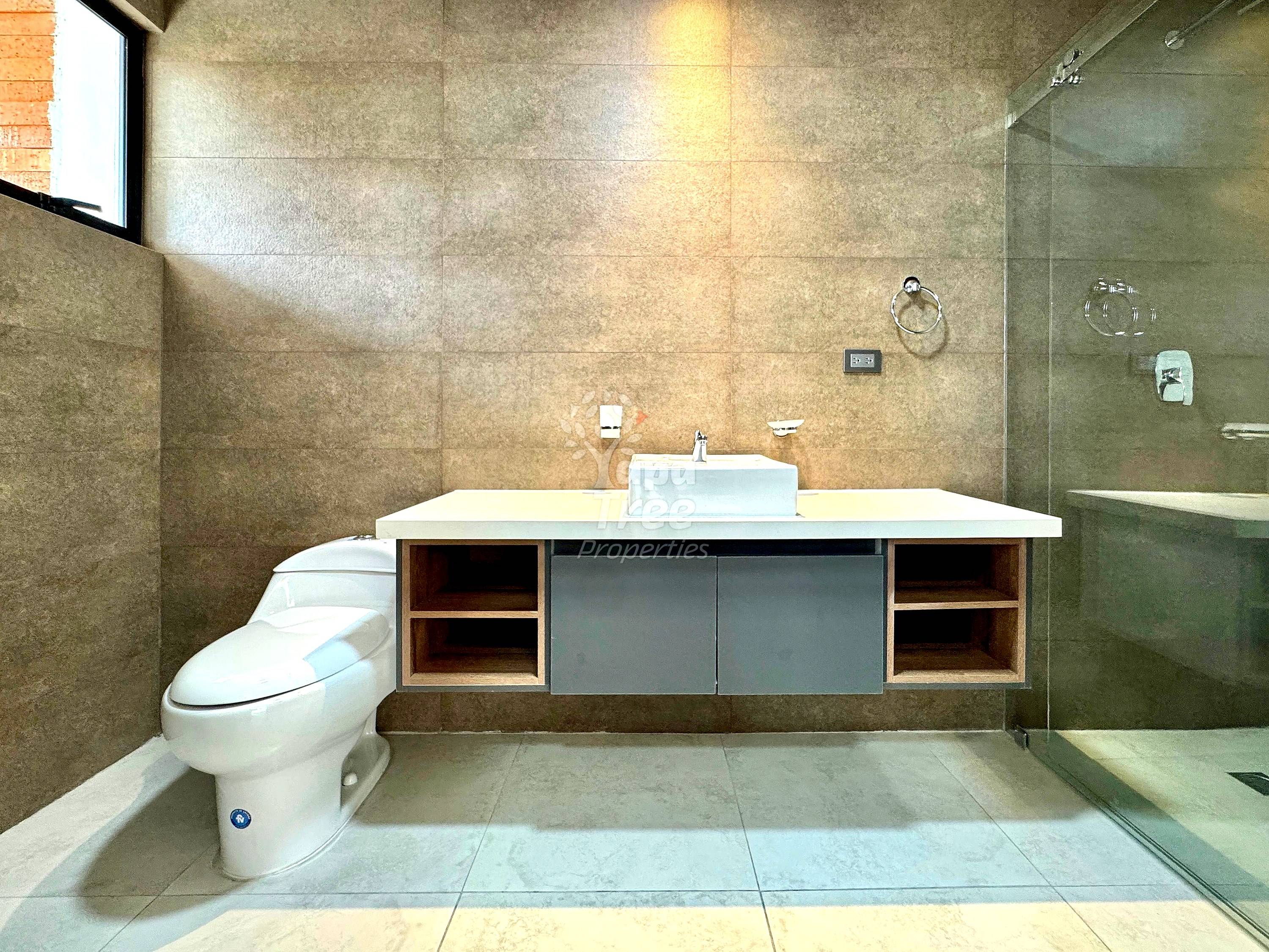 Modern bathroom with beige tile walls, floating vanity with white vessel sink, dark cabinetry with open shelving, toilet, glass shower enclosure, and window providing natural light.