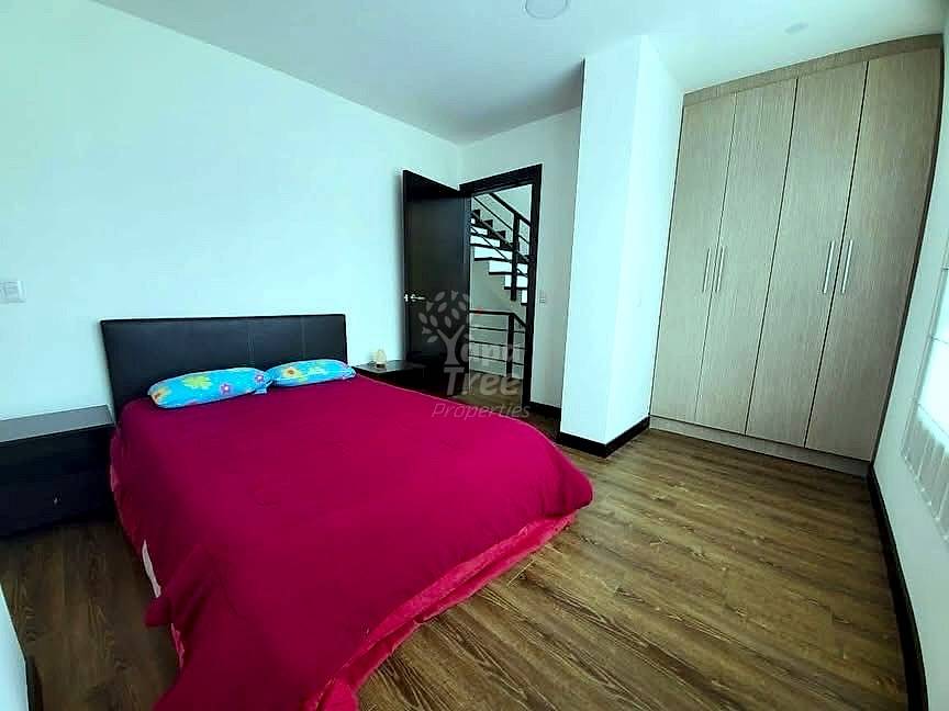 Bedroom with queen bed covered in red bedding, black headboard, wooden wardrobe, black metal door with stair view, wood flooring, white walls.