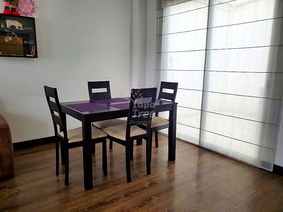 Dining room with black wooden table, four black chairs with cream cushions, wood-grain flooring, white walls, and sheer curtains over sliding glass doors.