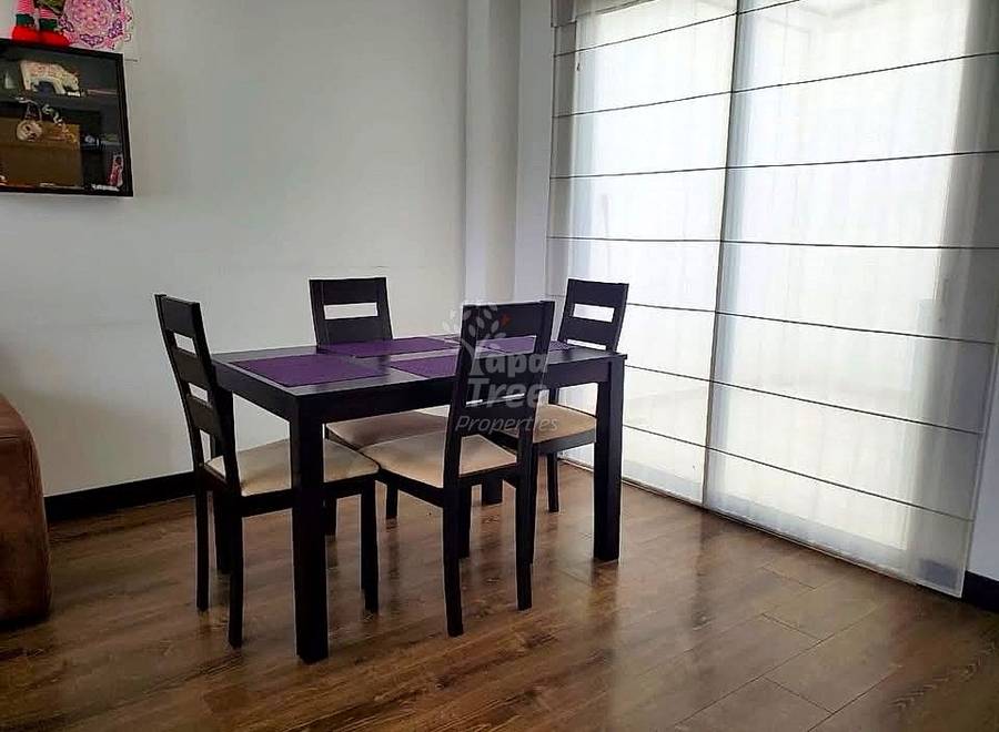Comedor con mesa de madera negra, cuatro sillas negras con cojines crema, piso de imitación madera, paredes blancas y cortinas translúcidas sobre puertas corredizas.