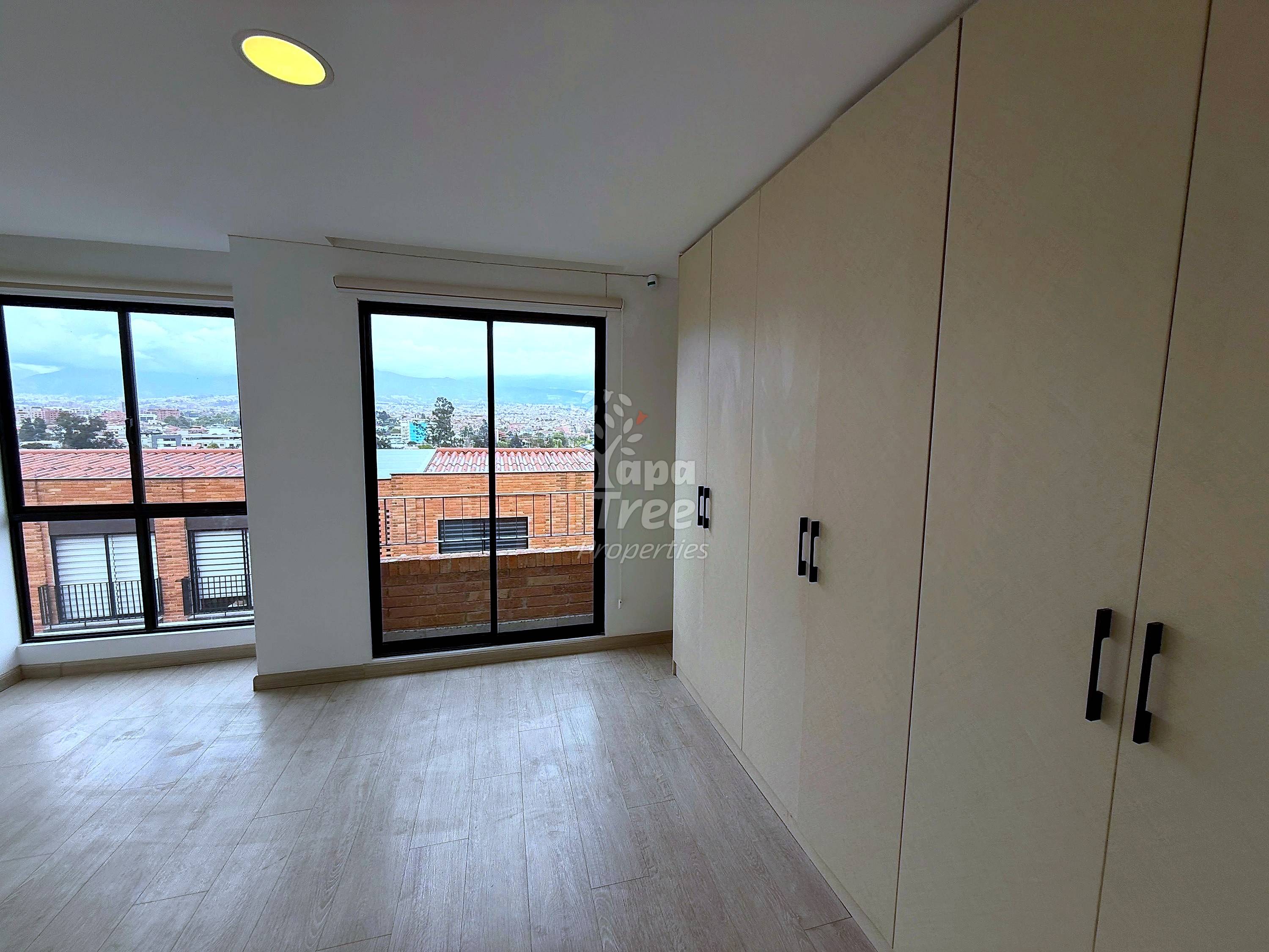 Bedroom with light wood flooring, beige built-in wardrobes with black handles, black-framed sliding glass doors and windows overlooking rooftops and Cuenca valley.