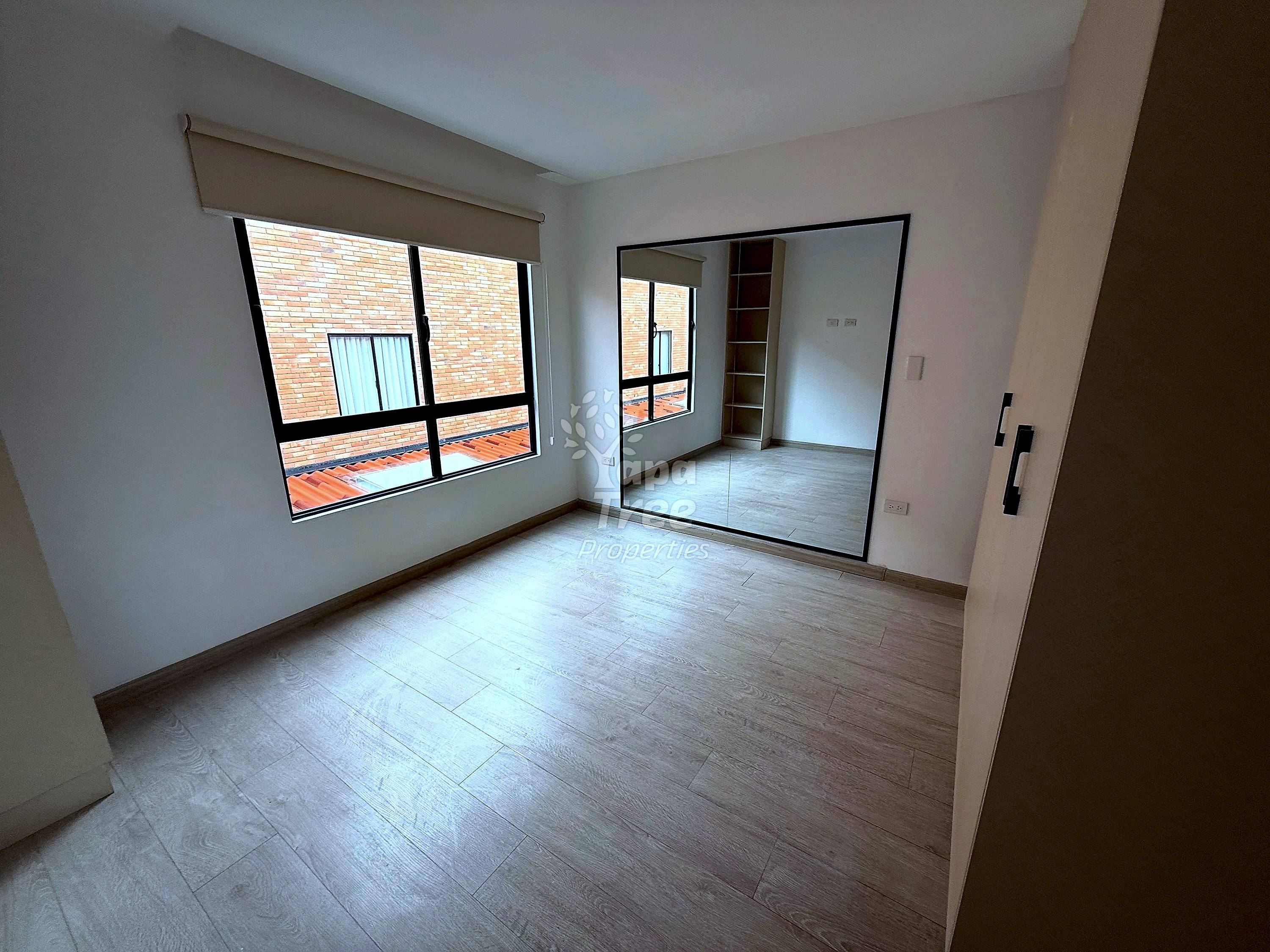 Empty bedroom with light wood flooring, large mirror, window with roller shade overlooking brick building, and bright natural lighting.