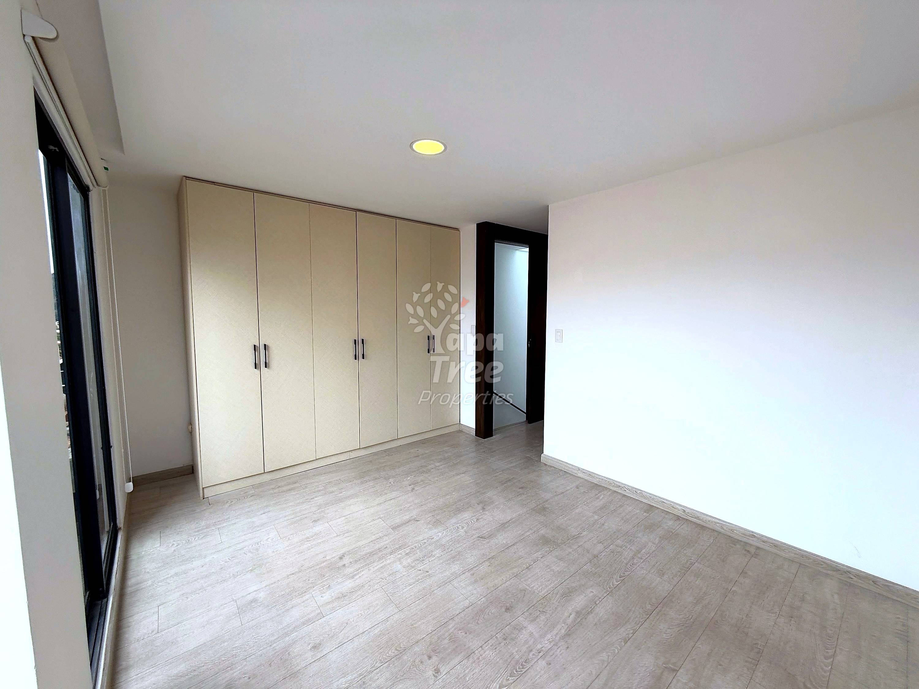 Bedroom with light wood laminate flooring, built-in beige wardrobe with six doors, recessed ceiling light, and doorway with dark frame.