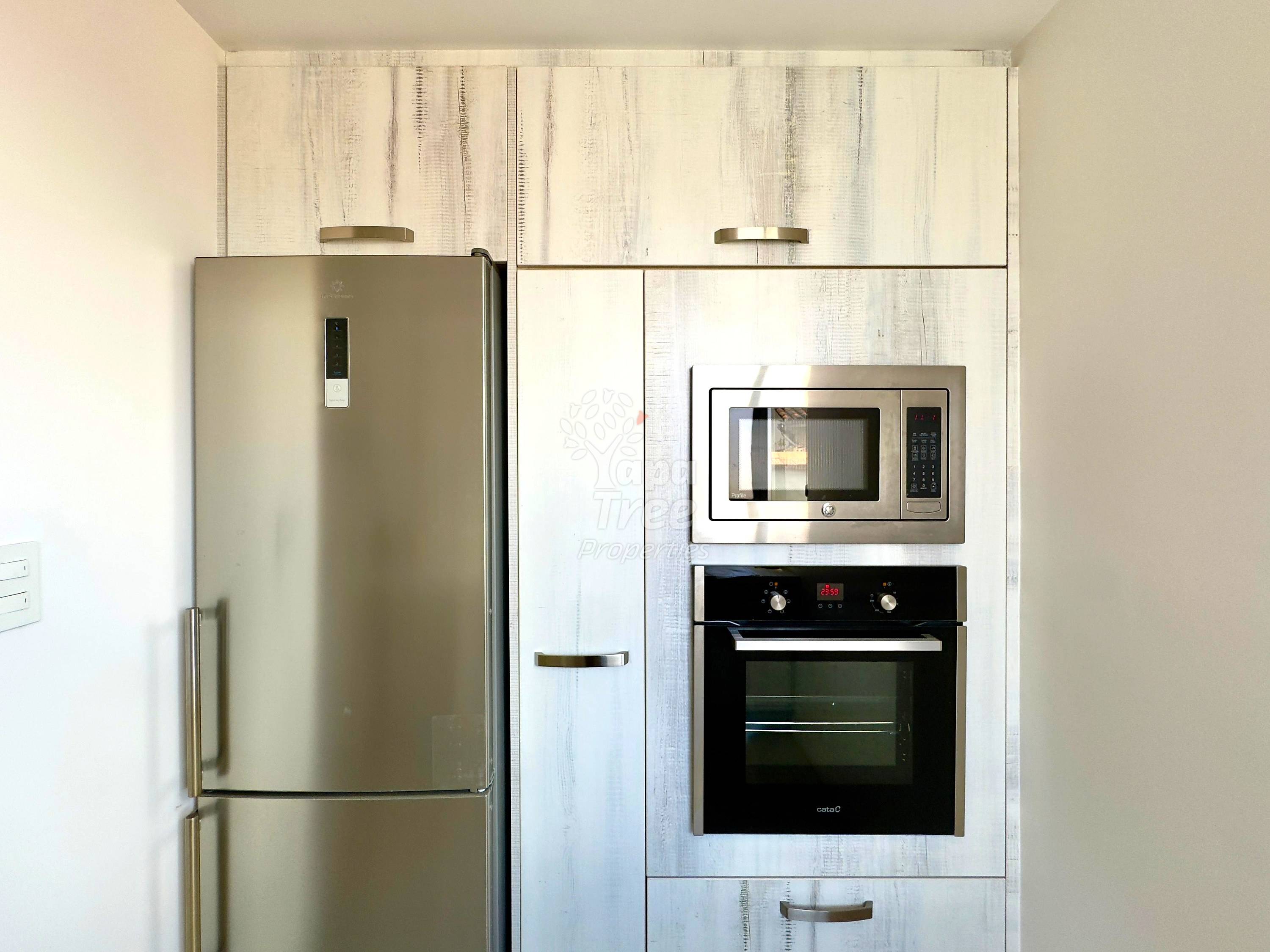 Kitchen with stainless steel refrigerator, white wood-grain cabinets, built-in microwave and oven, and metal hardware under bright overhead lighting.