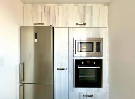 Kitchen with stainless steel refrigerator, white wood-grain cabinets, built-in microwave and oven, and metal hardware under bright overhead lighting.