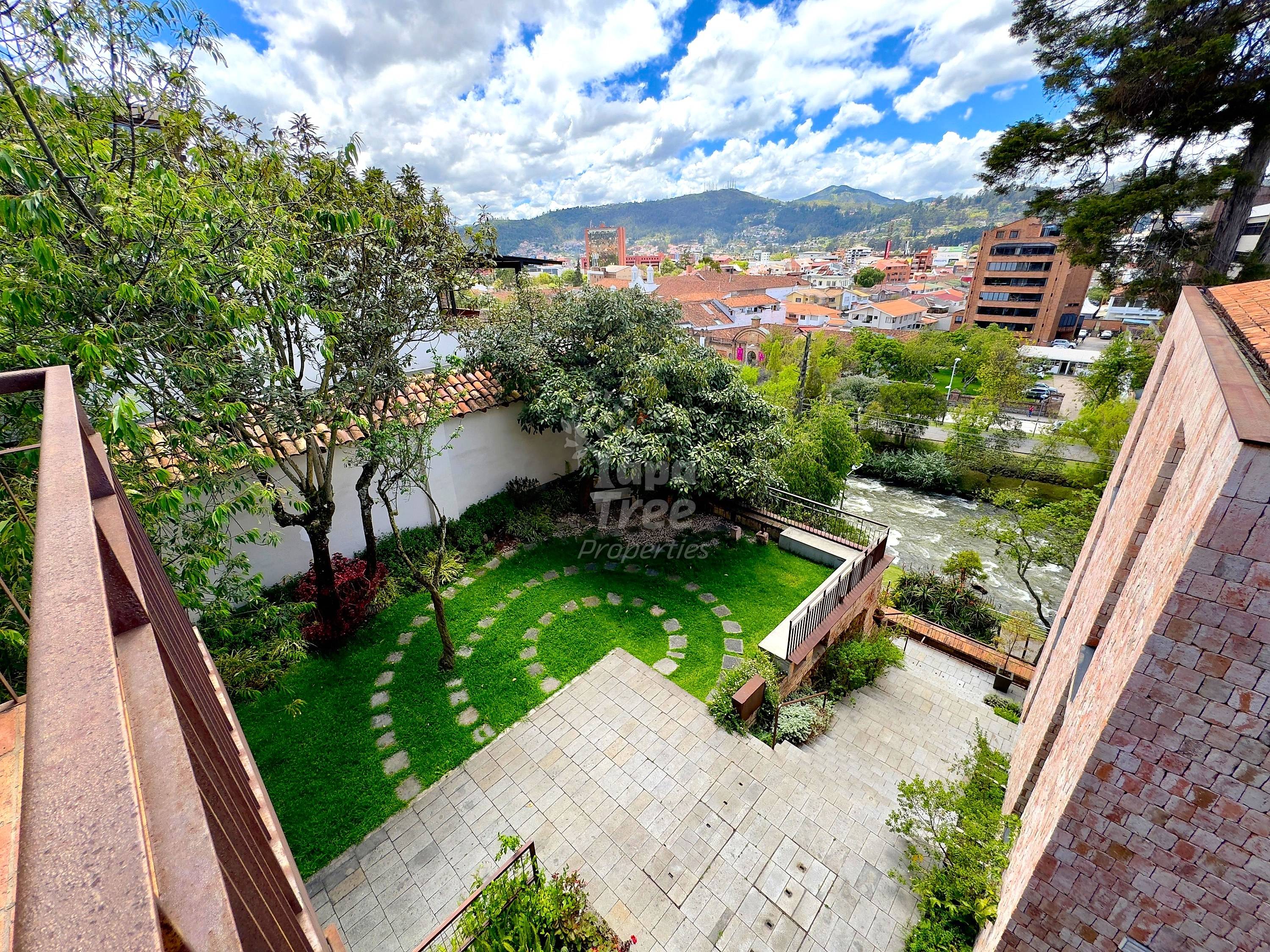 Overhead view of private courtyard with green lawn, stone pathway, trees, and brick boundary walls overlooking Cuenca cityscape and river.