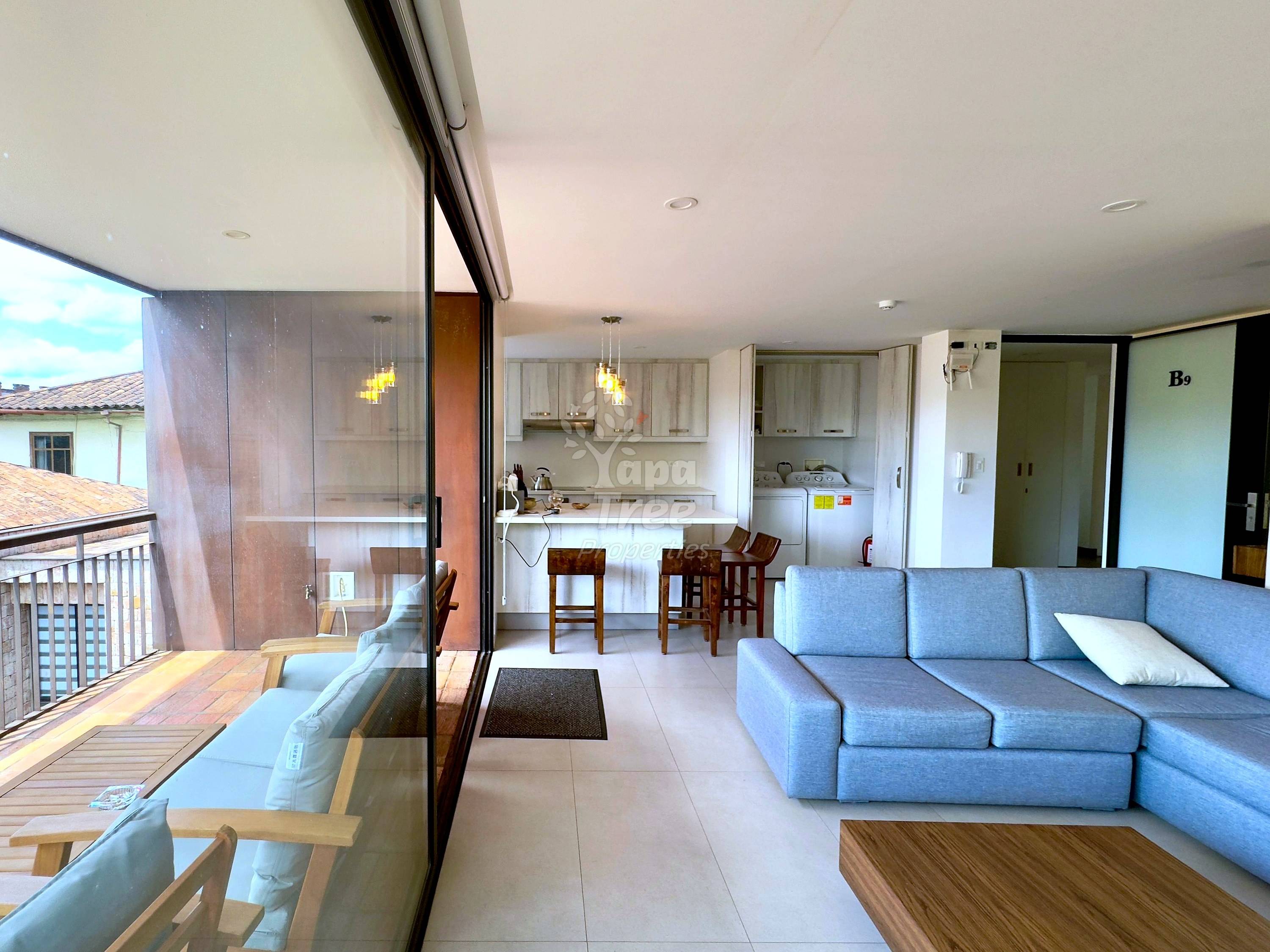 Open-plan departamento interior showing gray sectional sofa, wooden coffee table, dining area with wooden chairs, white cocina with cabinets, and glass door opening to balcony with city views.