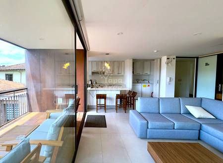 Open-plan departamento interior showing gray sectional sofa, wooden coffee table, dining area with wooden chairs, white cocina with cabinets, and glass door opening to balcony with city views.