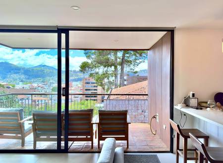 Interior room with sliding glass doors opening to terrace, wooden chairs, desk with monitor, mountain and city views of Cuenca visible beyond railing.