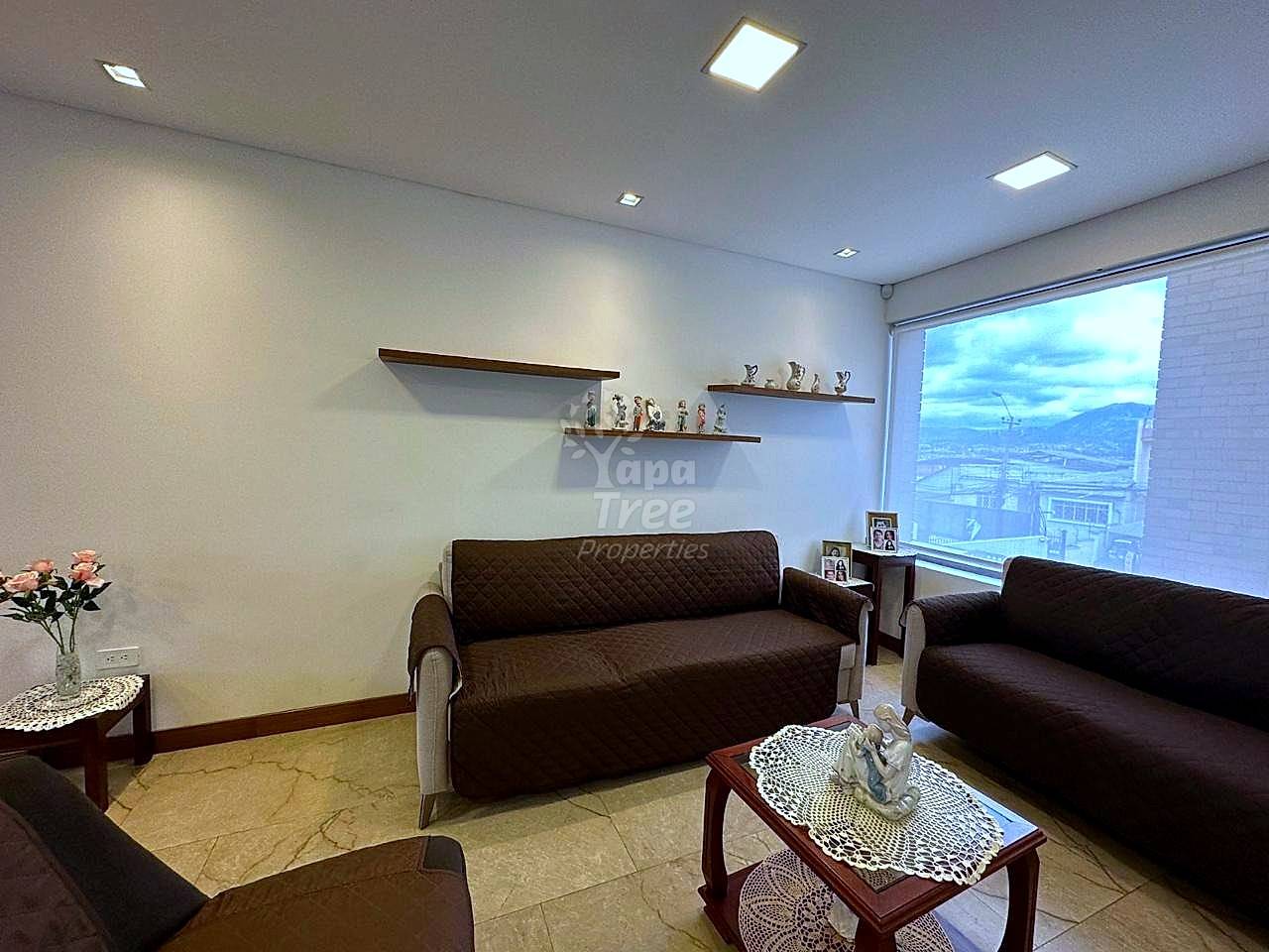 Living room with two dark brown sofas, marble floor, wooden floating shelves, recessed ceiling lights, and large window with city views.