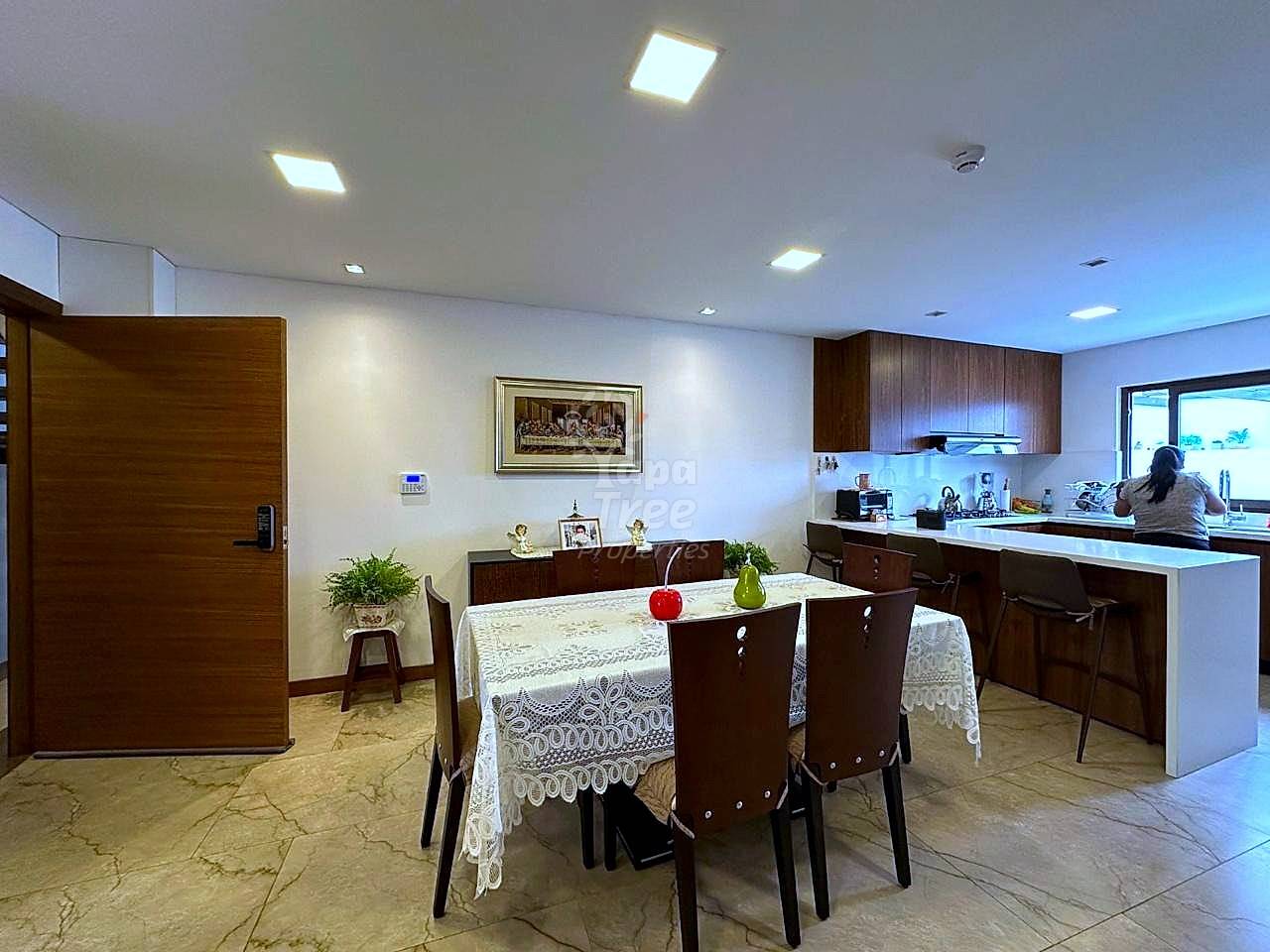 Open-plan departamento with dining table, dark wood chairs, white countertop kitchen, recessed ceiling lights, marble tile flooring, and brown wood entry door.