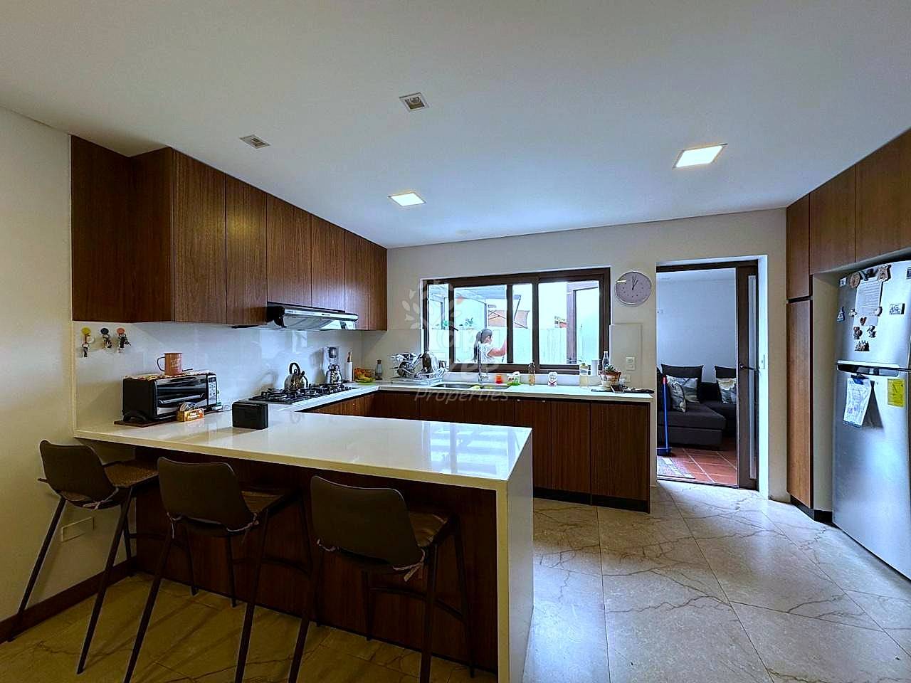 Kitchen with dark wood cabinets, white countertop island with seating, stainless steel refrigerator, recessed ceiling lights, and tile flooring throughout.